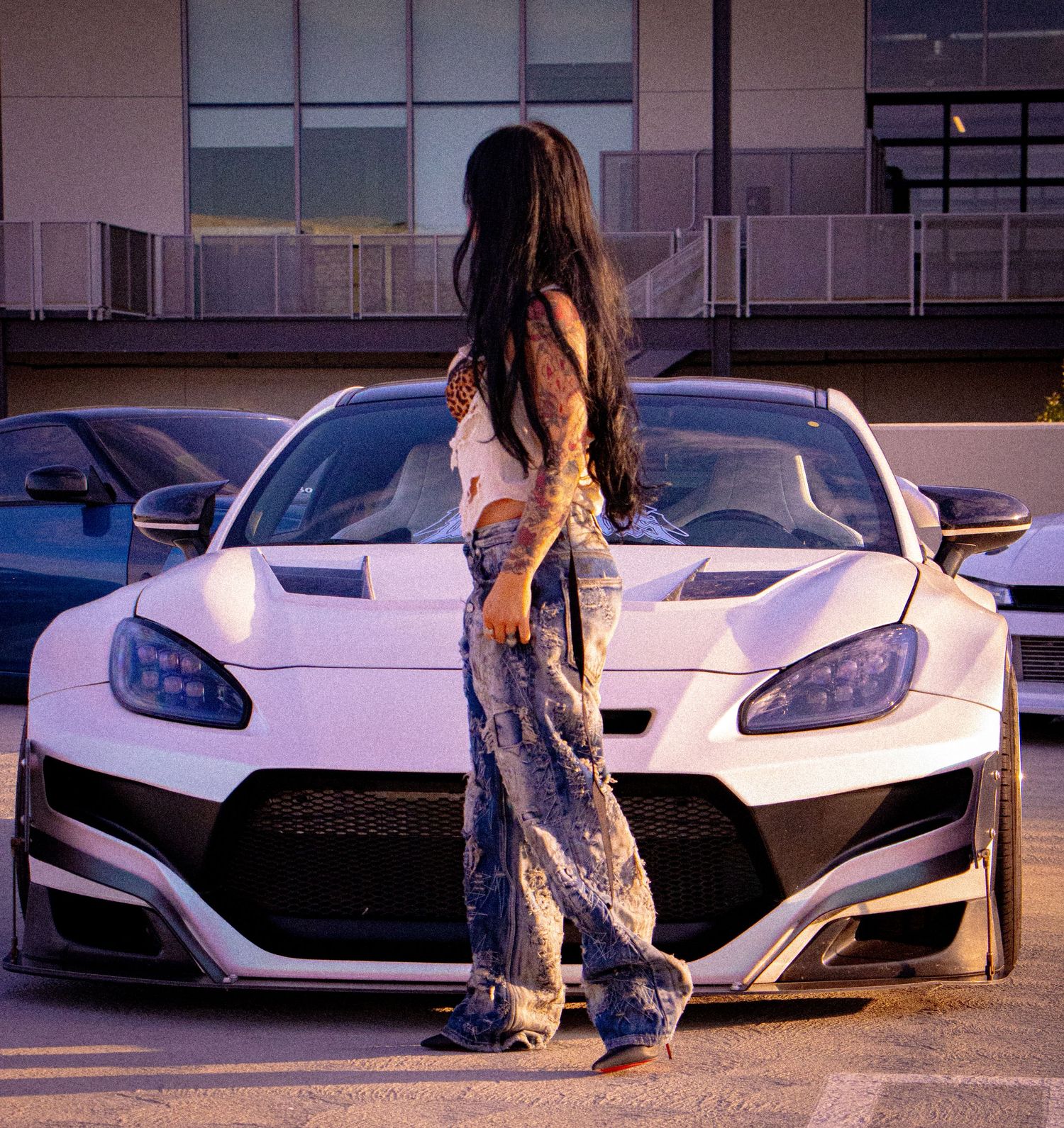 Tattooed woman in jeans poses in front of a white widebody sports car in a parking lot at dusk.