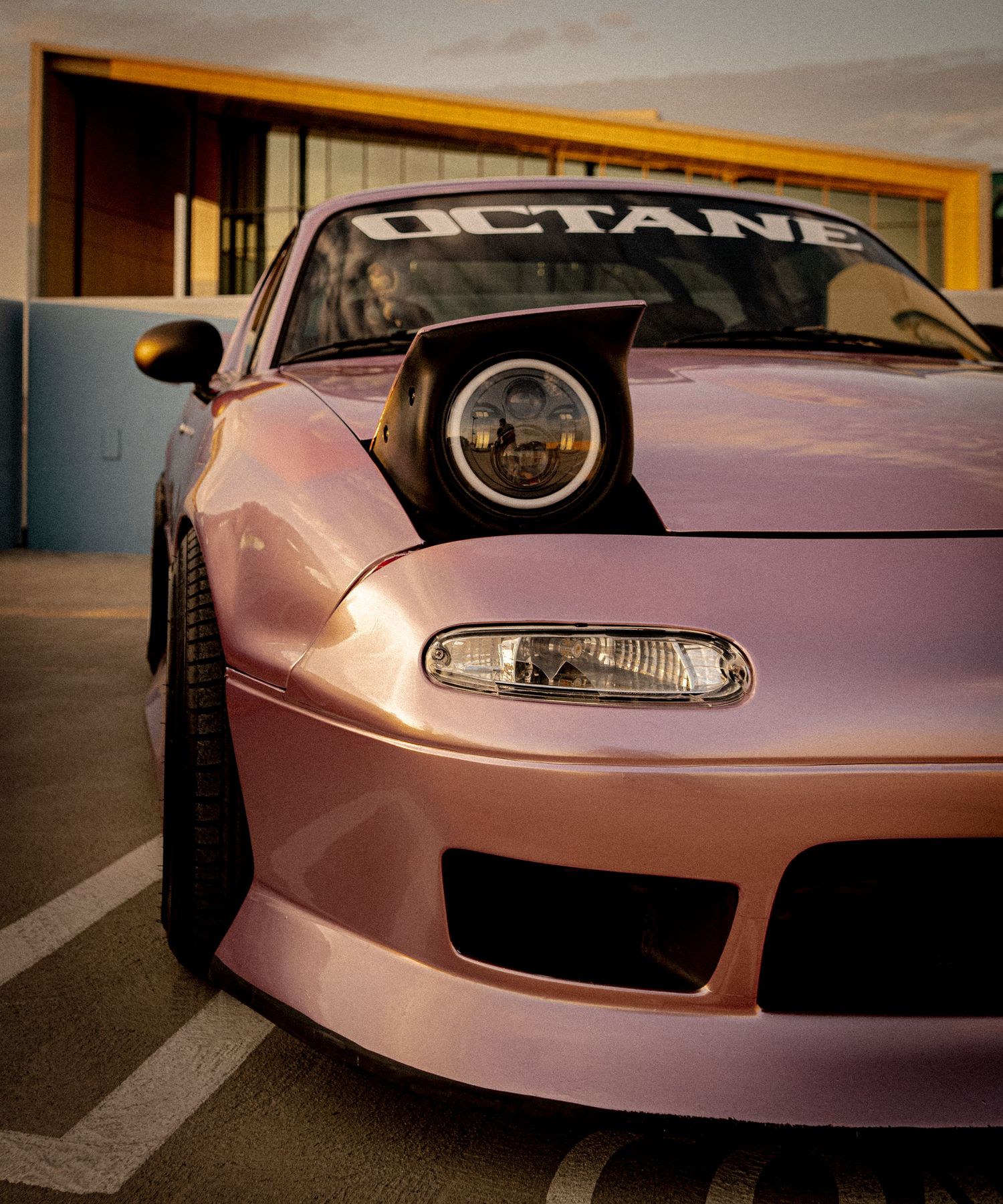 Pink modified sports car with OCTANE windshield banner and pop-up headlight parked outside a building at sunset.