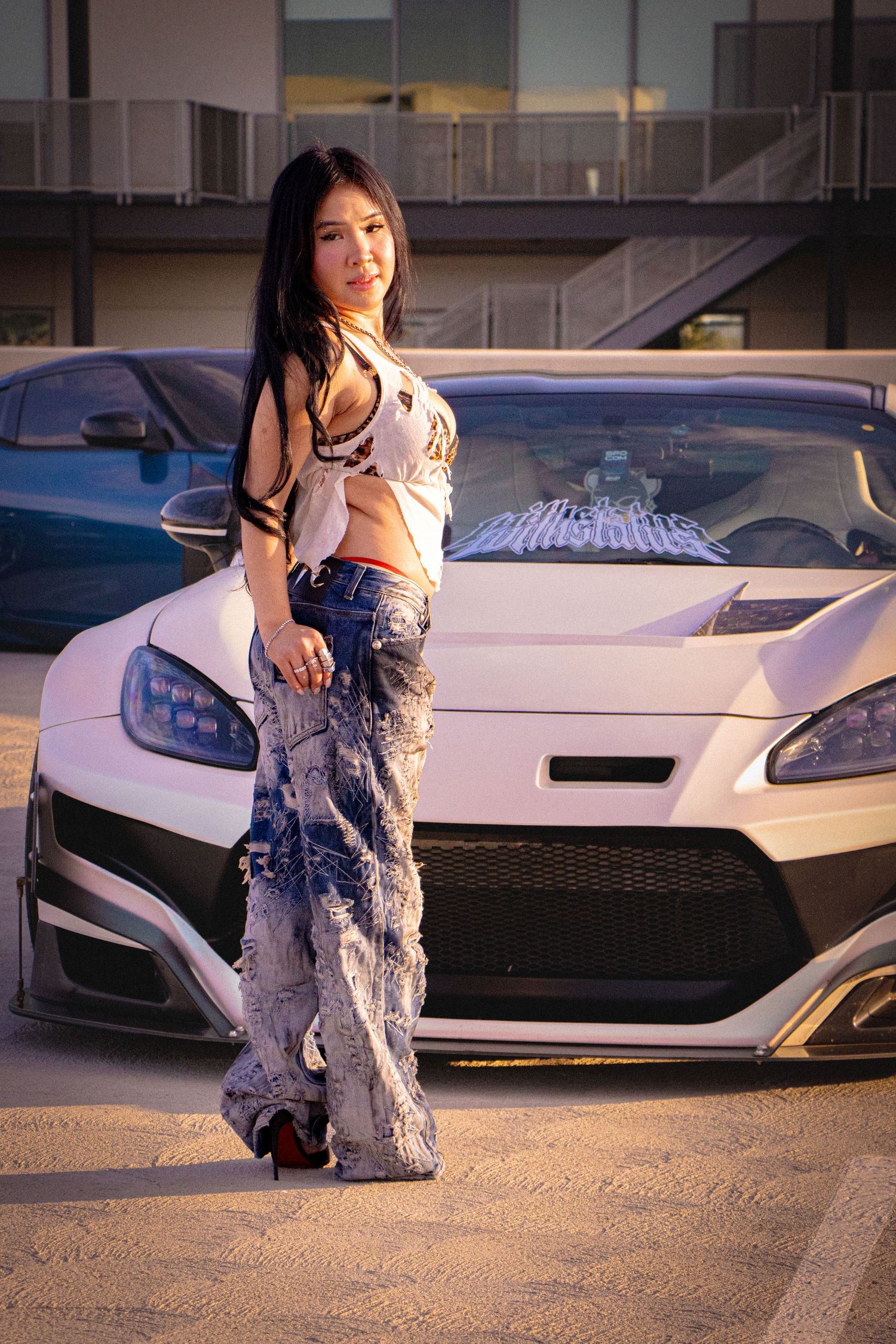 Young woman in crop top and wide-leg jeans poses confidently against a white sports car in a parking lot at sunset.