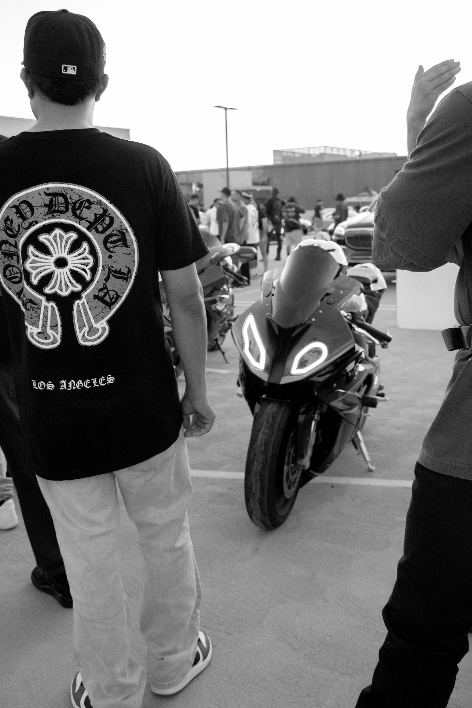 Black and white photo of a man in Chrome Hearts LA shirt standing near a sport motorcycle at an outdoor gathering.