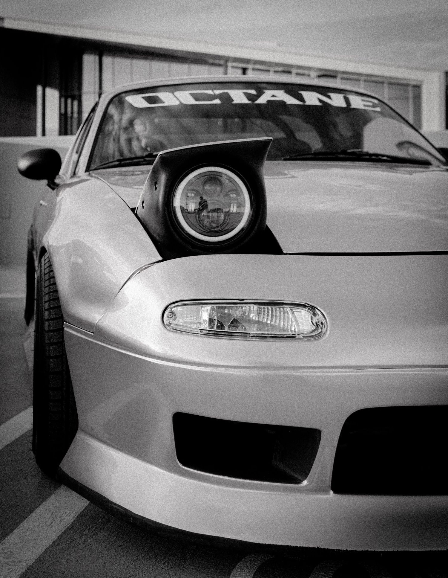 Black and white photo of a Mazda Miata with pop-up headlight and Octane windshield banner at a car meet.