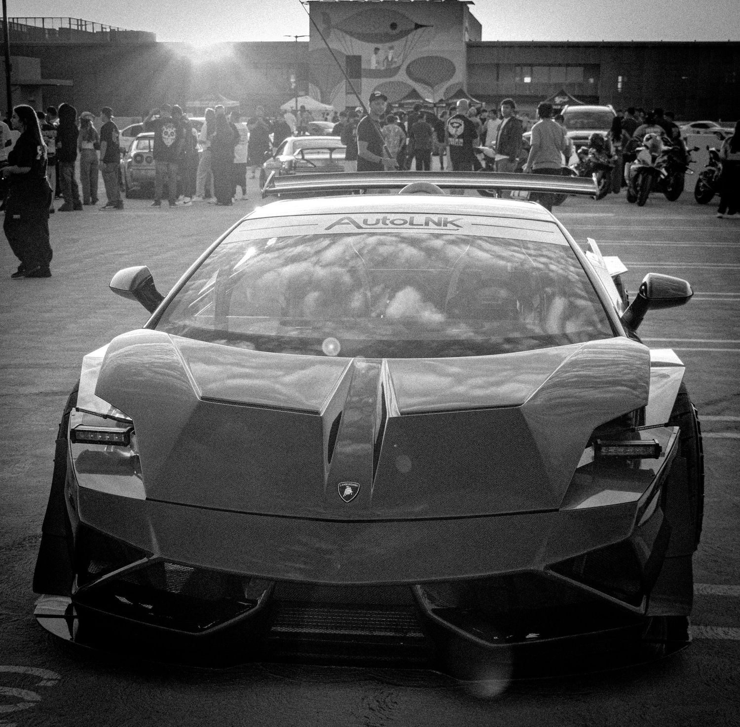 Black and white photo of a Lamborghini Aventador with AutoLink branding at an outdoor car meet.