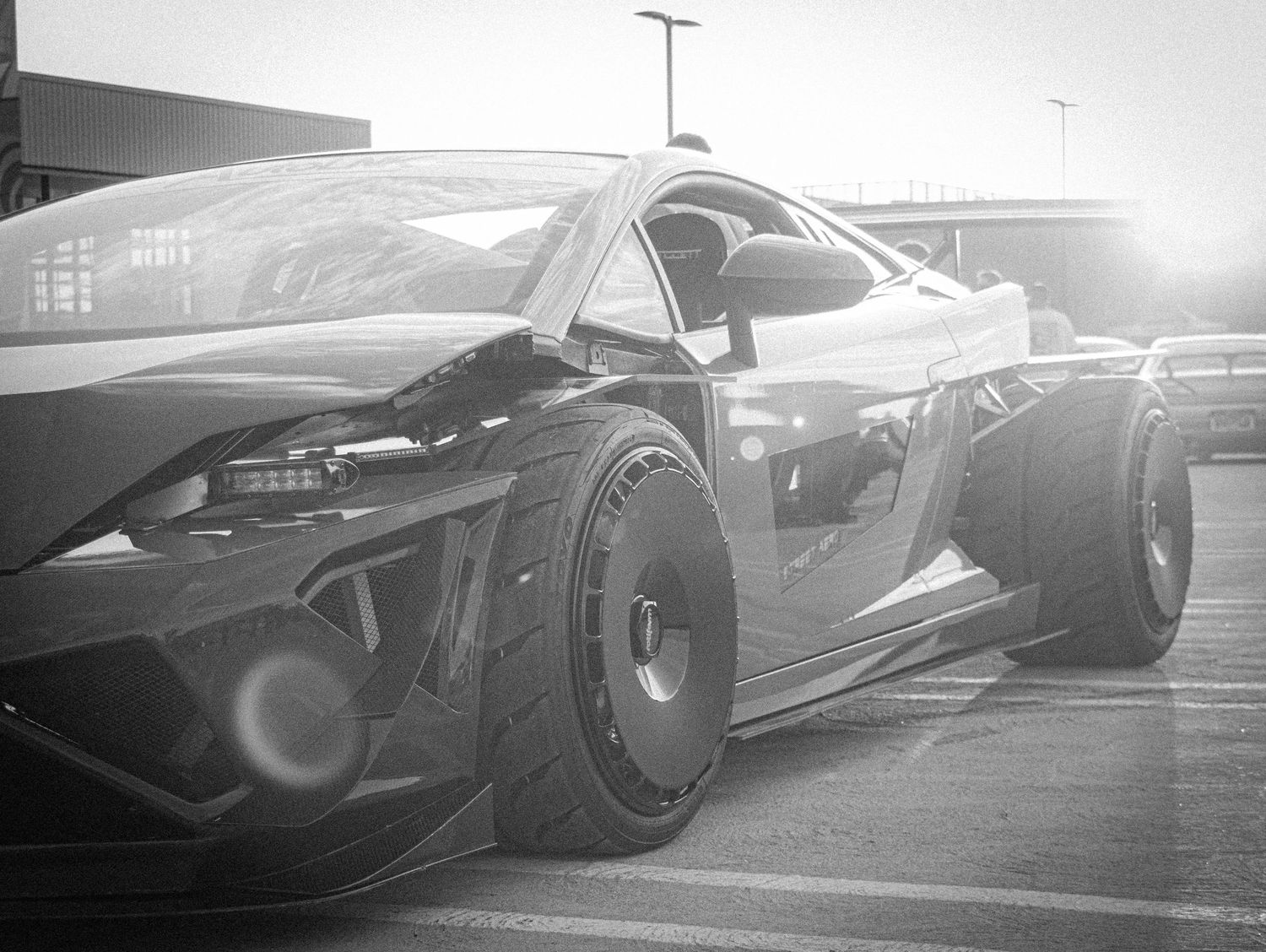 Black and white photo of a futuristic custom supercar with wide body kit and oversized wheels in a parking lot.