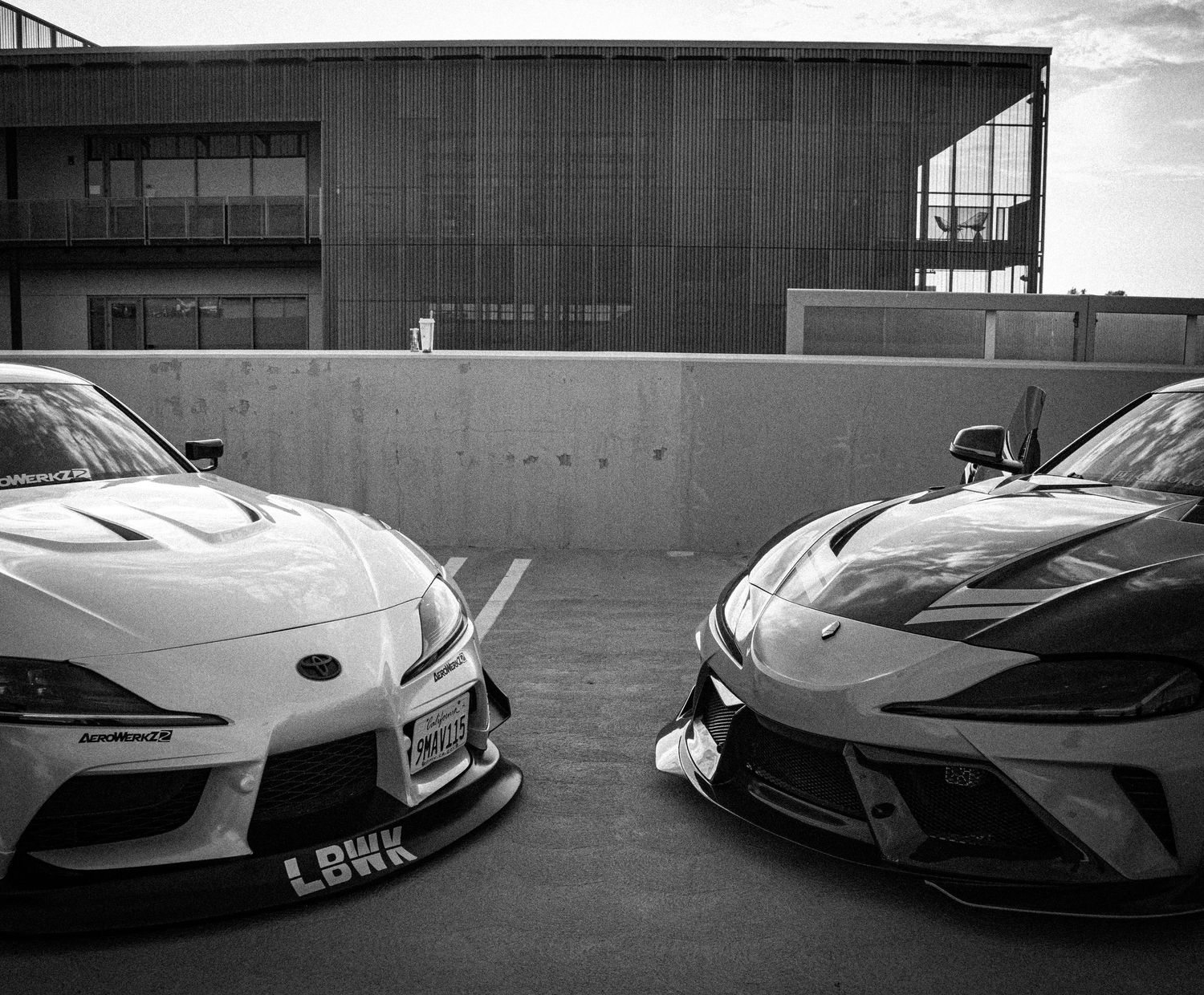Two modified sports cars, a Toyota Supra and Corvette with body kits, parked side by side in a black and white photo.
