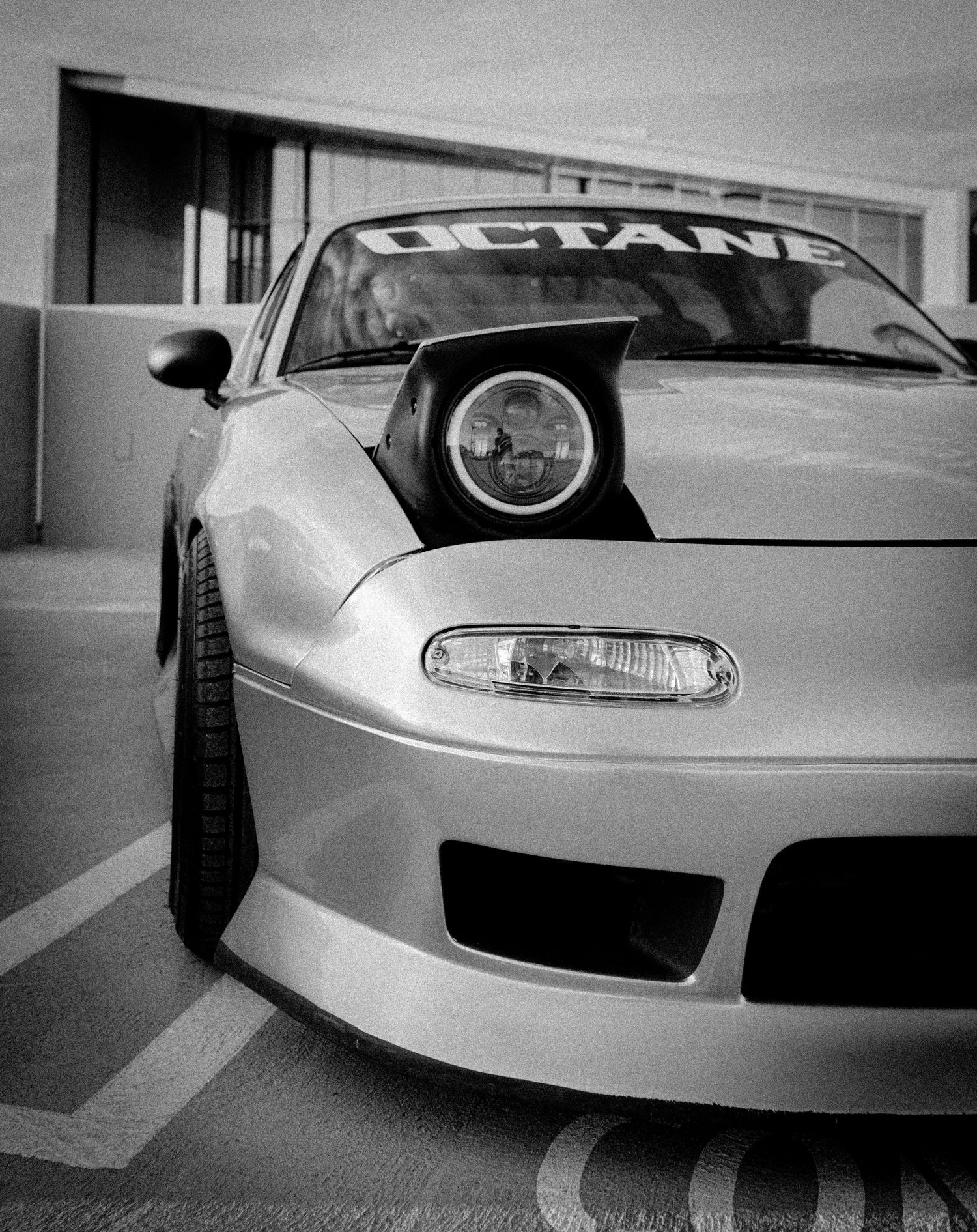 Black and white front view of a modified Mazda RX-7 with OCTAN windshield banner parked in a lot.