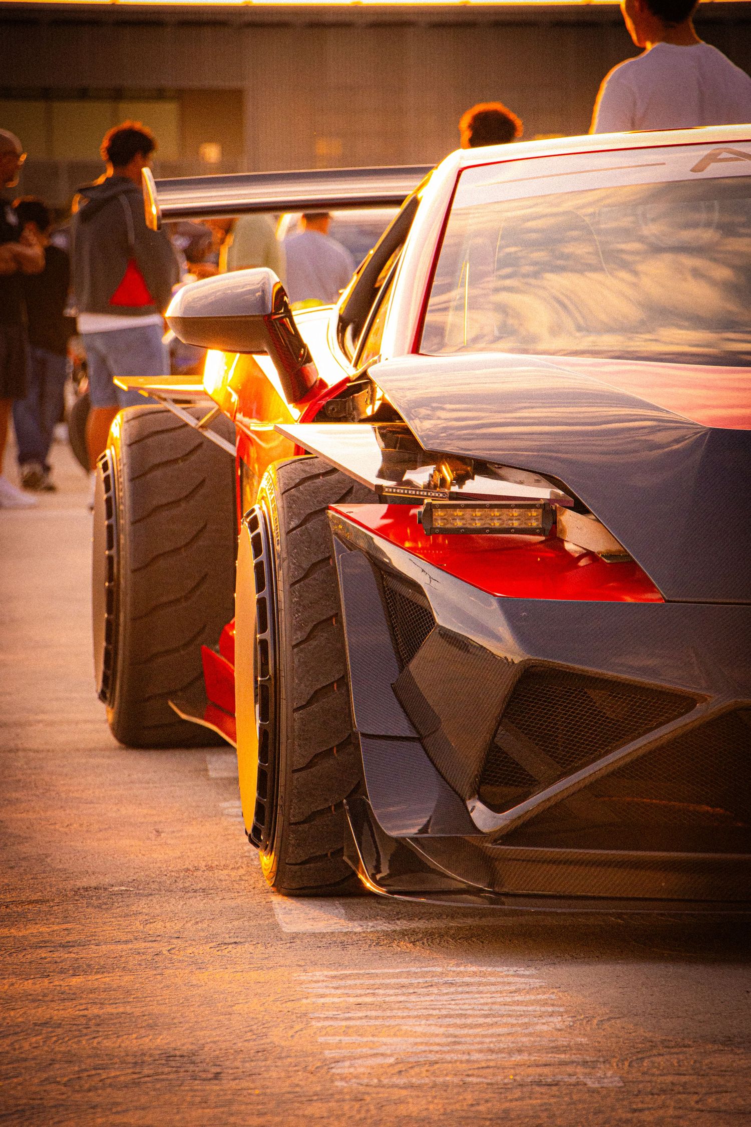 A sleek black Lamborghini supercar glows under golden sunset light at an outdoor car show with spectators in the background.