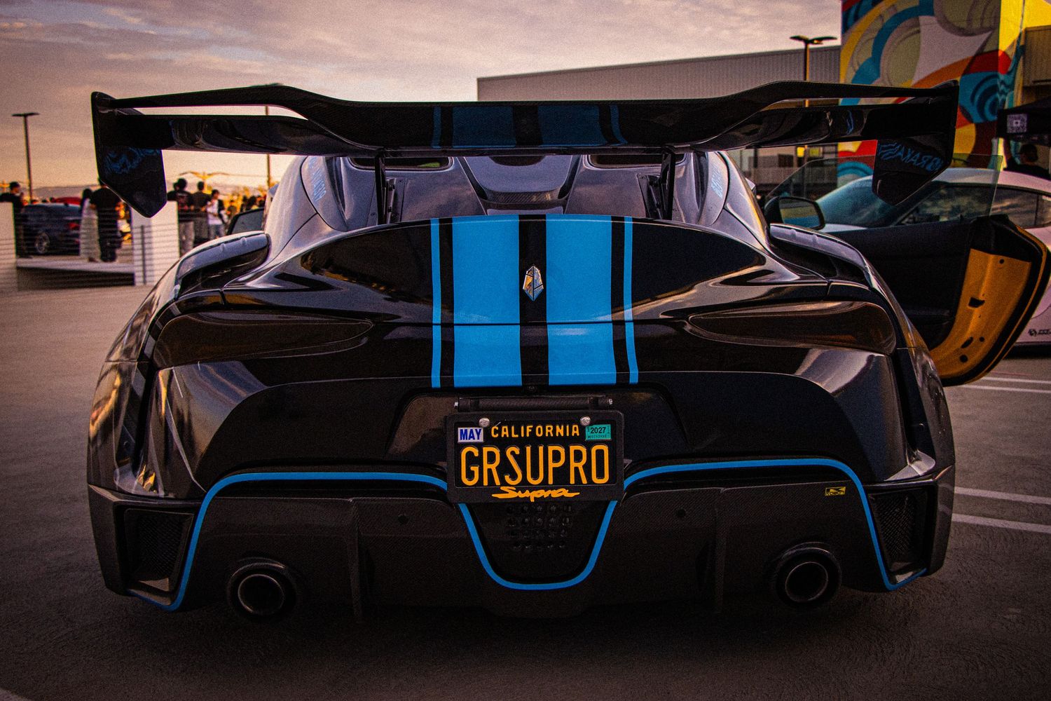 Black supercar with blue racing stripes and GRSUPRO California license plate photographed from rear at sunset.