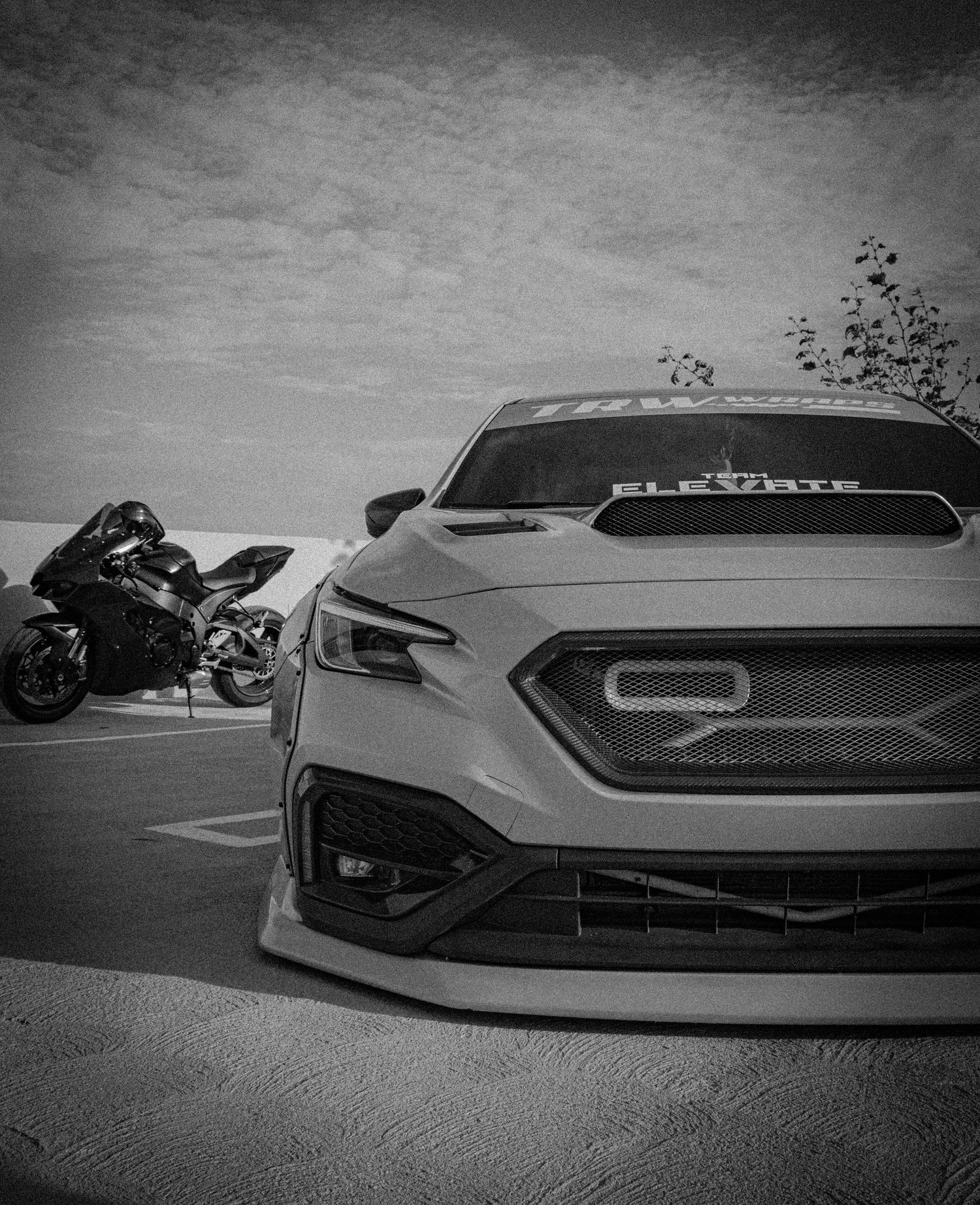 Black and white photo of a modified sports car with aggressive front bumper and a motorcycle parked beside it.
