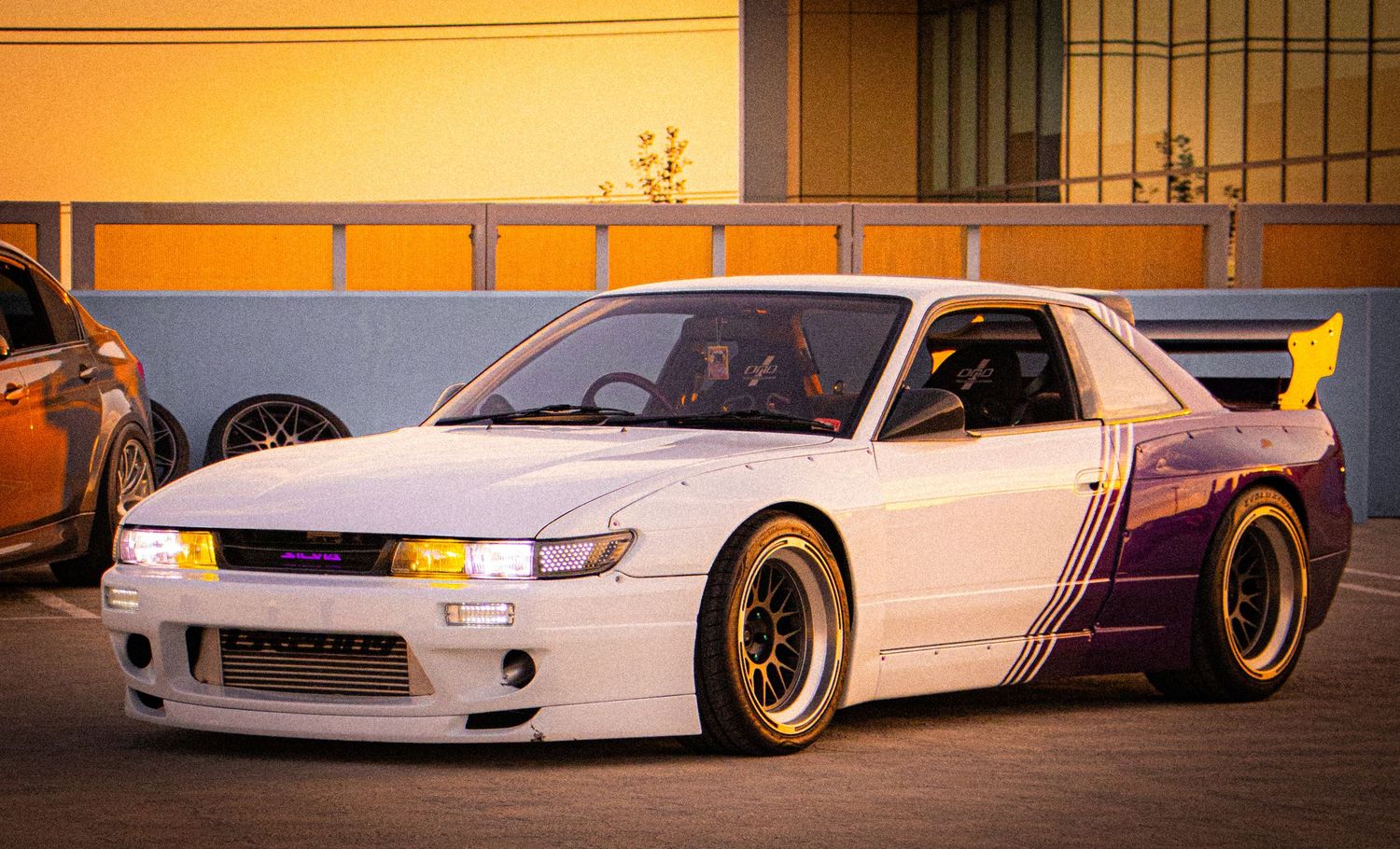 Modified white Nissan Silvia S13 with purple accents, wide body kit, and gold wheels at a car meet.