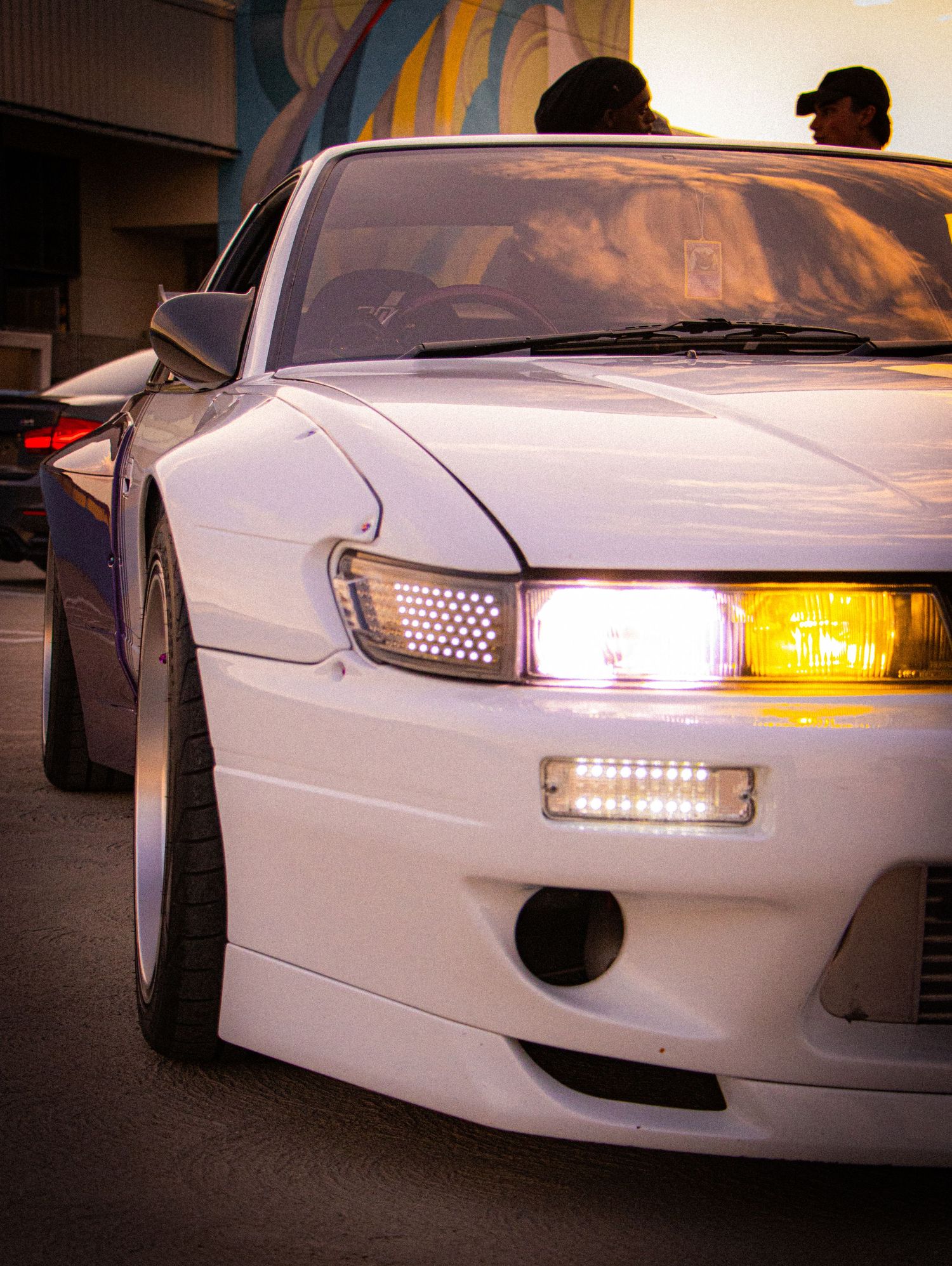 White Nissan Silvia S13 with yellow headlights and LED DRL at a car meet, low stance and wide body kit.
