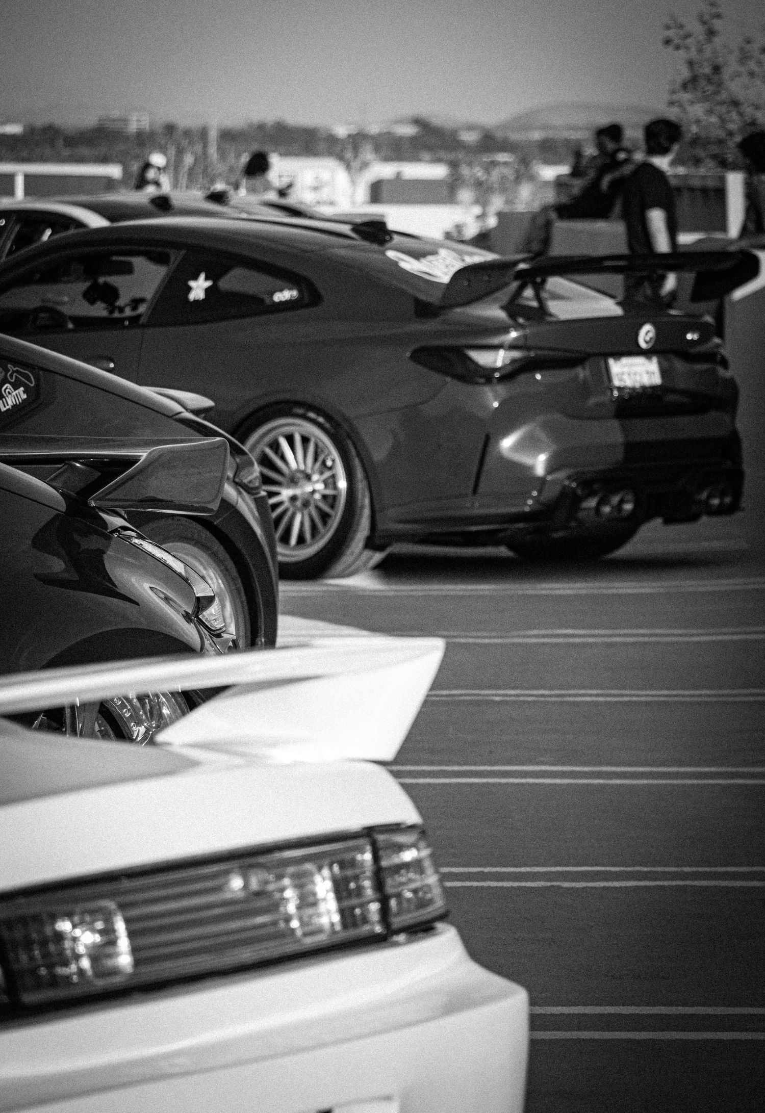 Black and white photo of modified sports cars, including a Toyota Supra with rear spoiler, at an outdoor car meet.