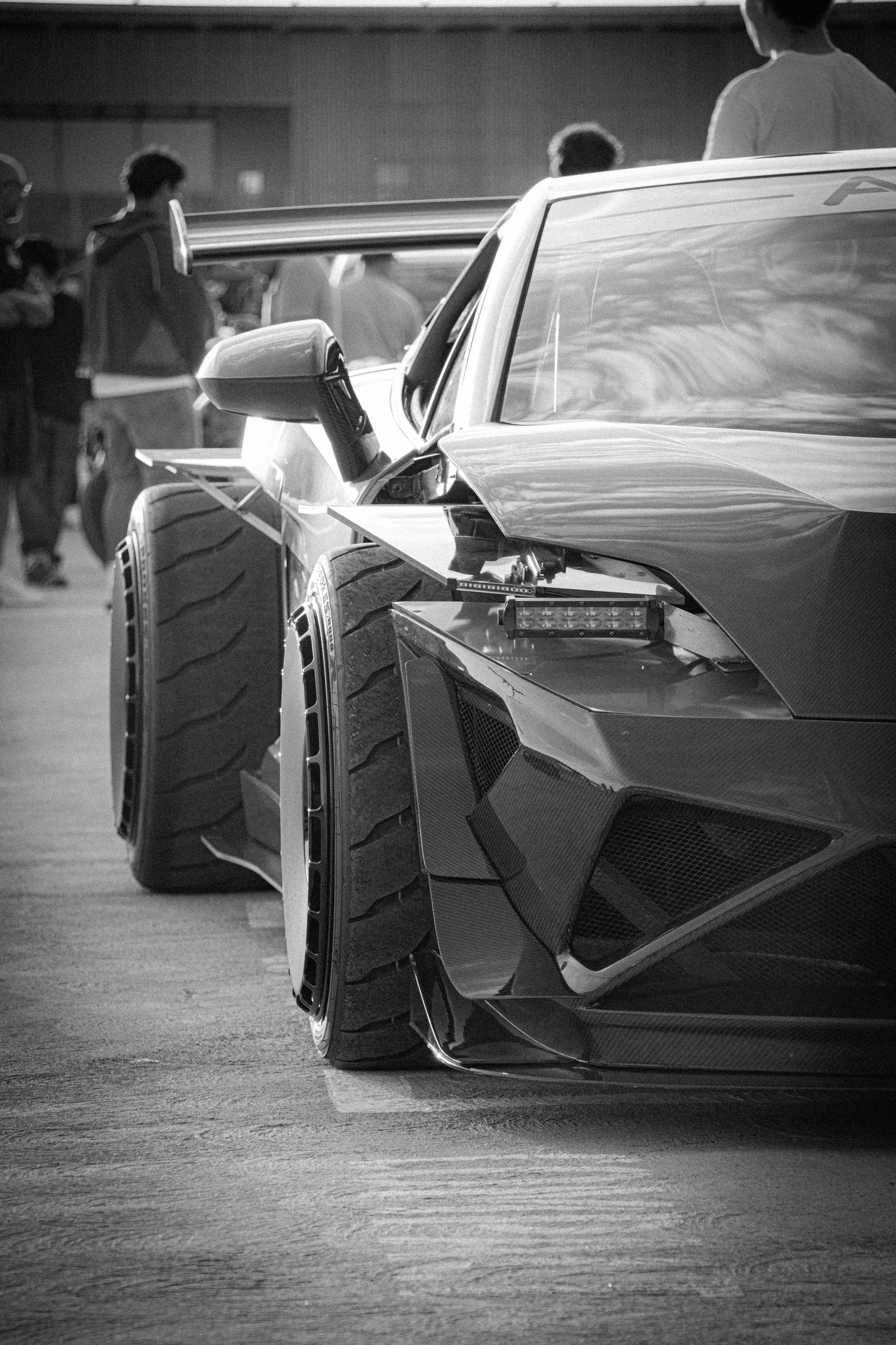 Black and white photo of a sleek Lamborghini supercar with wide body kit and large rear wing at a car show.