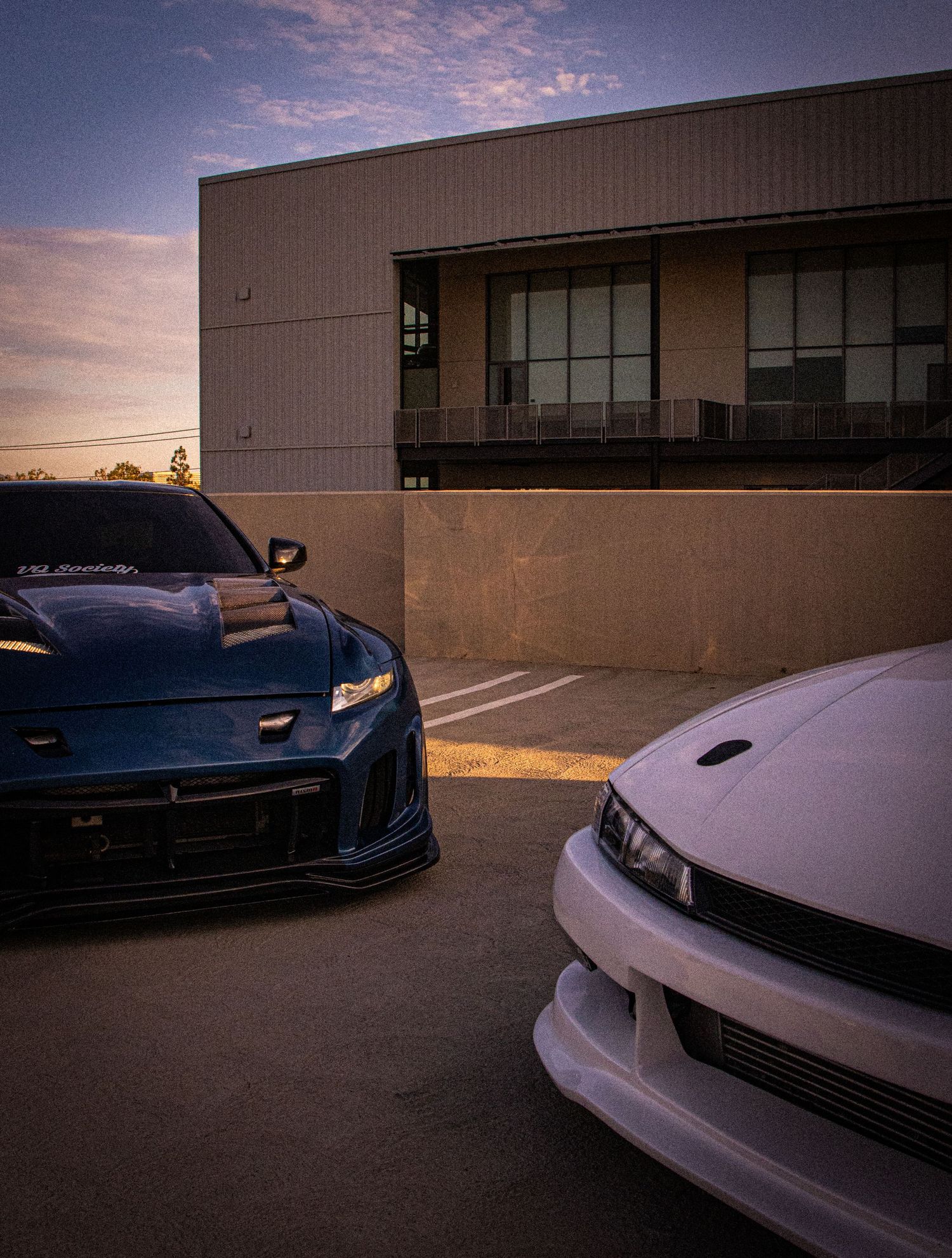 Two modified sports cars, a blue and white, parked at sunset near a modern building.