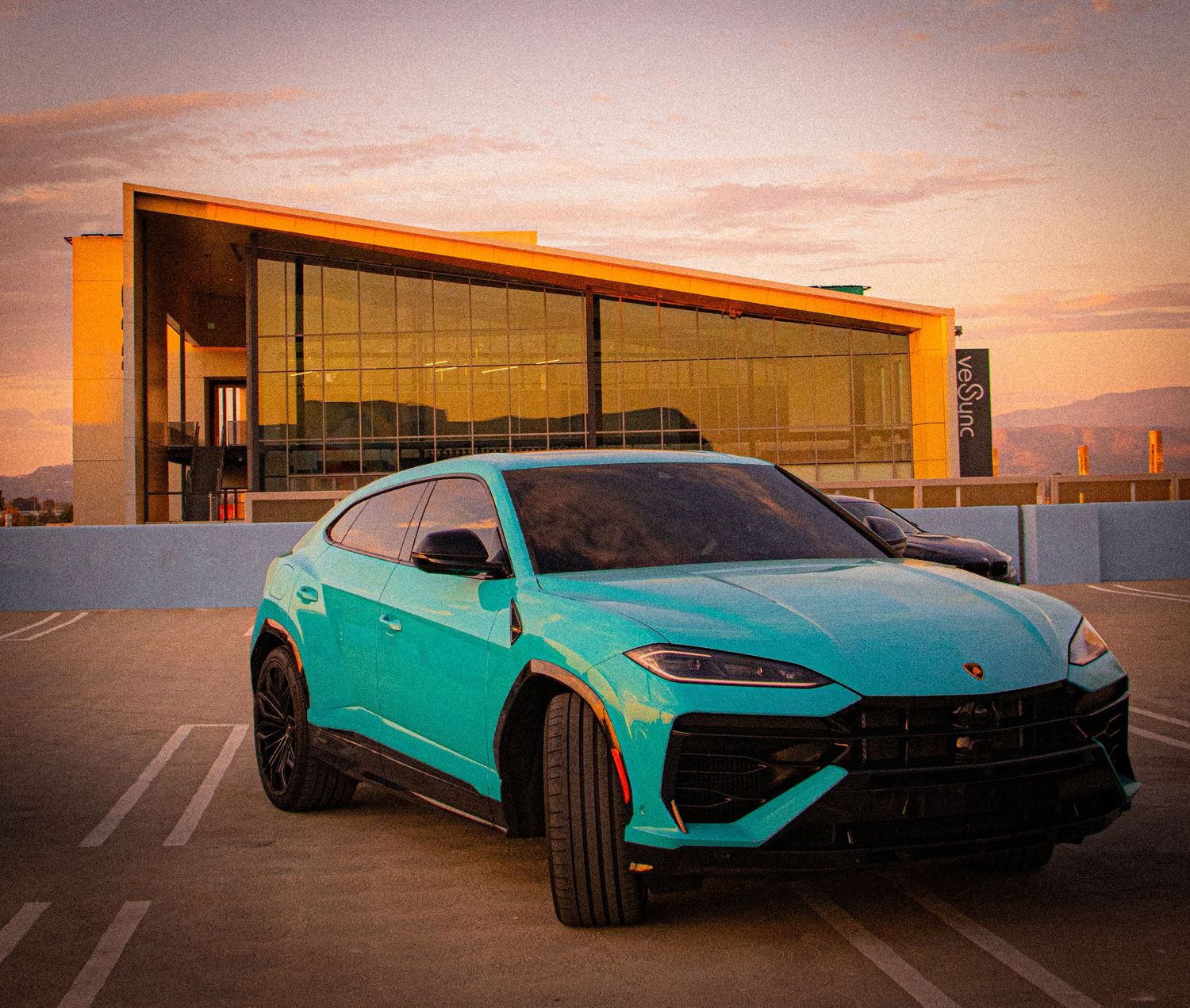 Teal Lamborghini Urus SUV parked on rooftop lot at golden sunset with modern glass building backdrop.