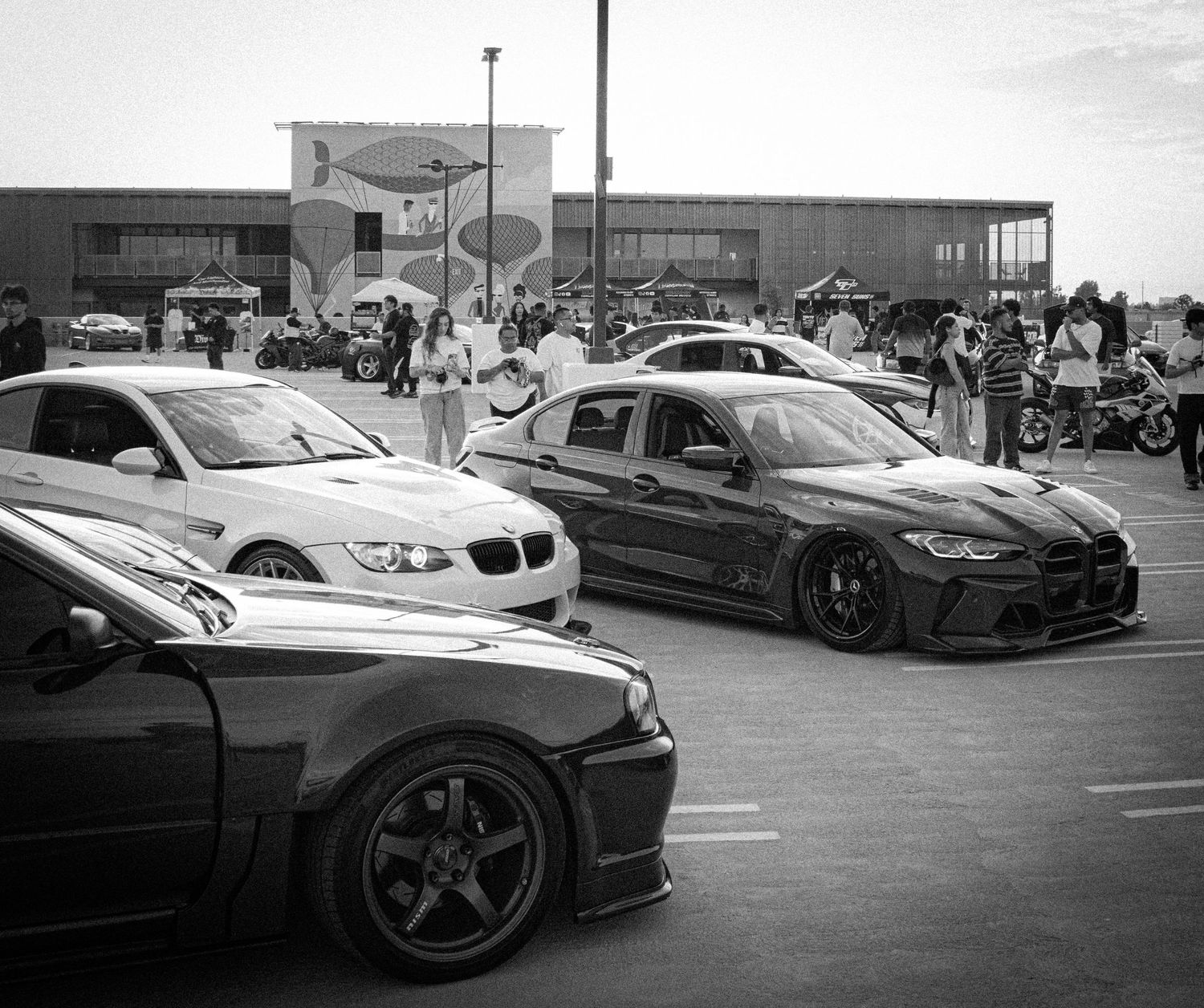 Black and white photo of modified BMW cars parked at an outdoor car show event with spectators.