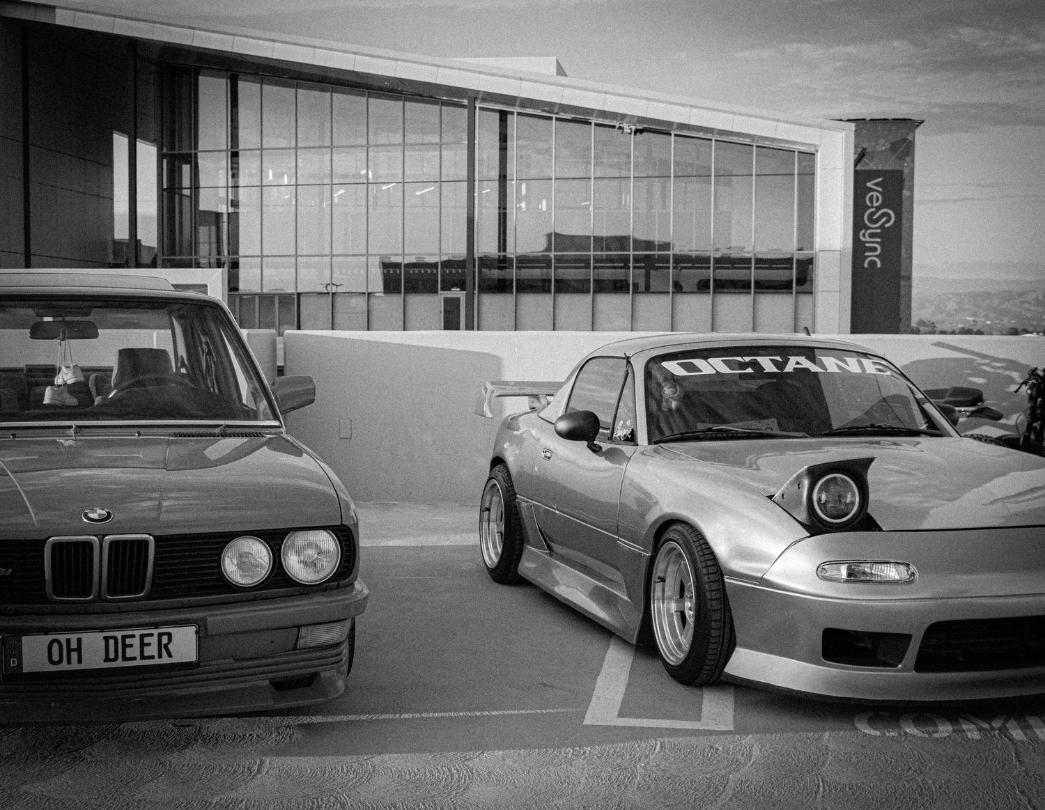 Black and white photo of two modified cars, a BMW E28 and Mazda RX-7, parked outside a modern building.