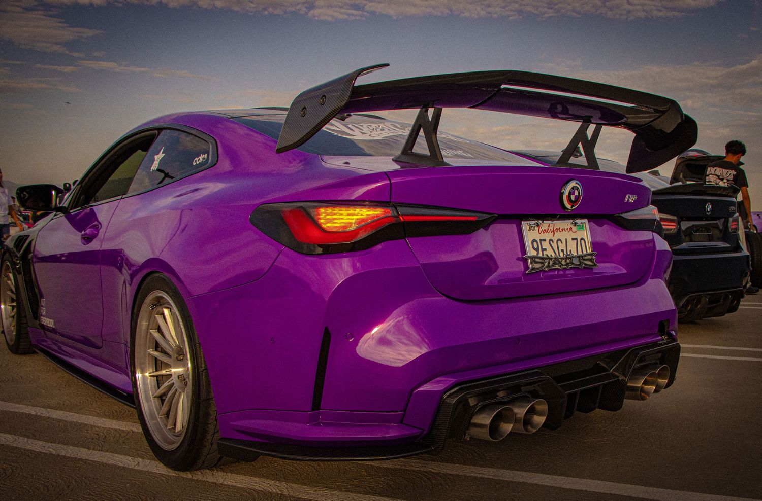 Vibrant purple BMW coupe with large rear spoiler and carbon fiber diffuser parked at a car show at dusk.
