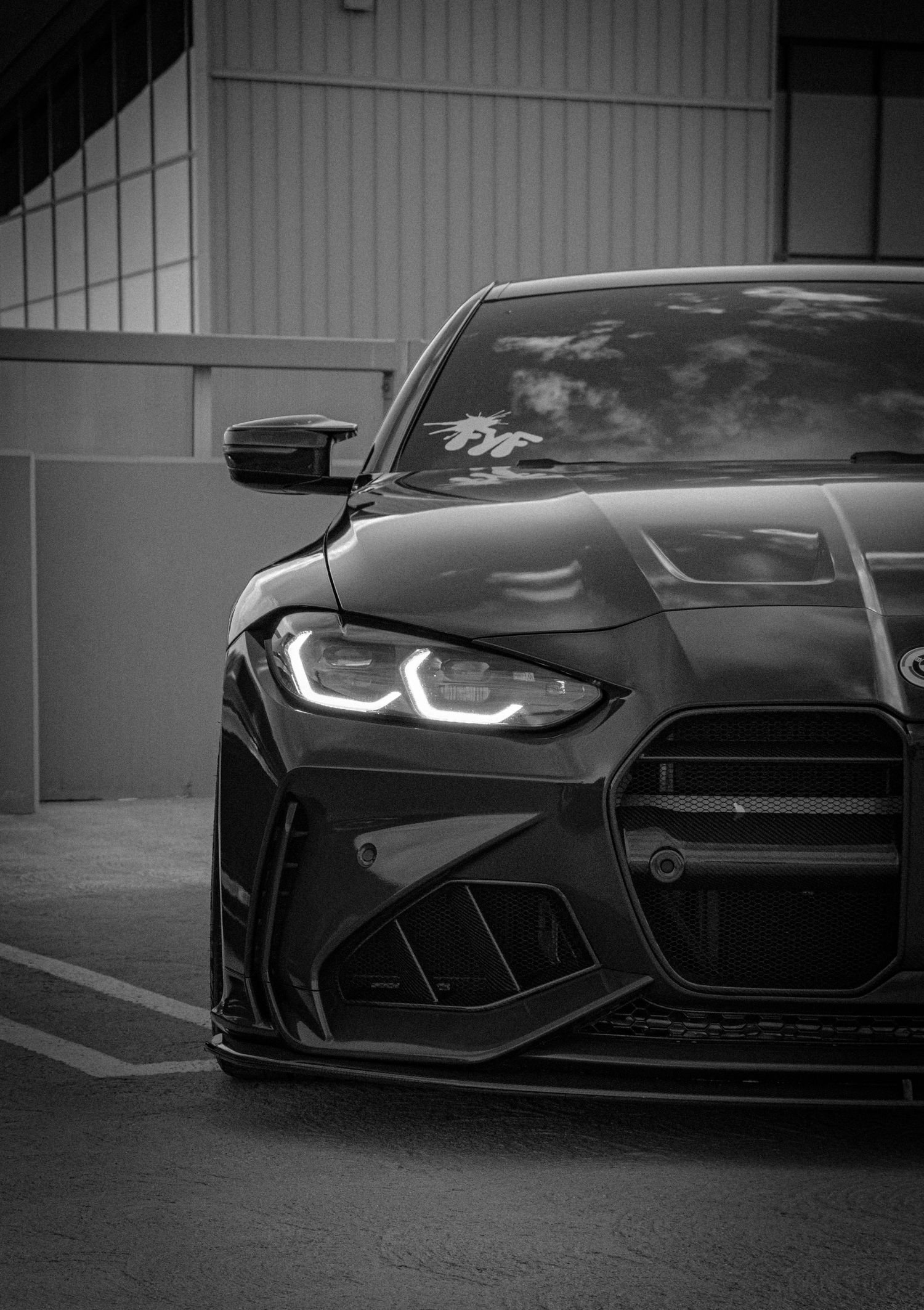 Black BMW M4 with aggressive wide body kit and LED headlights captured in dramatic black and white front-facing photo.