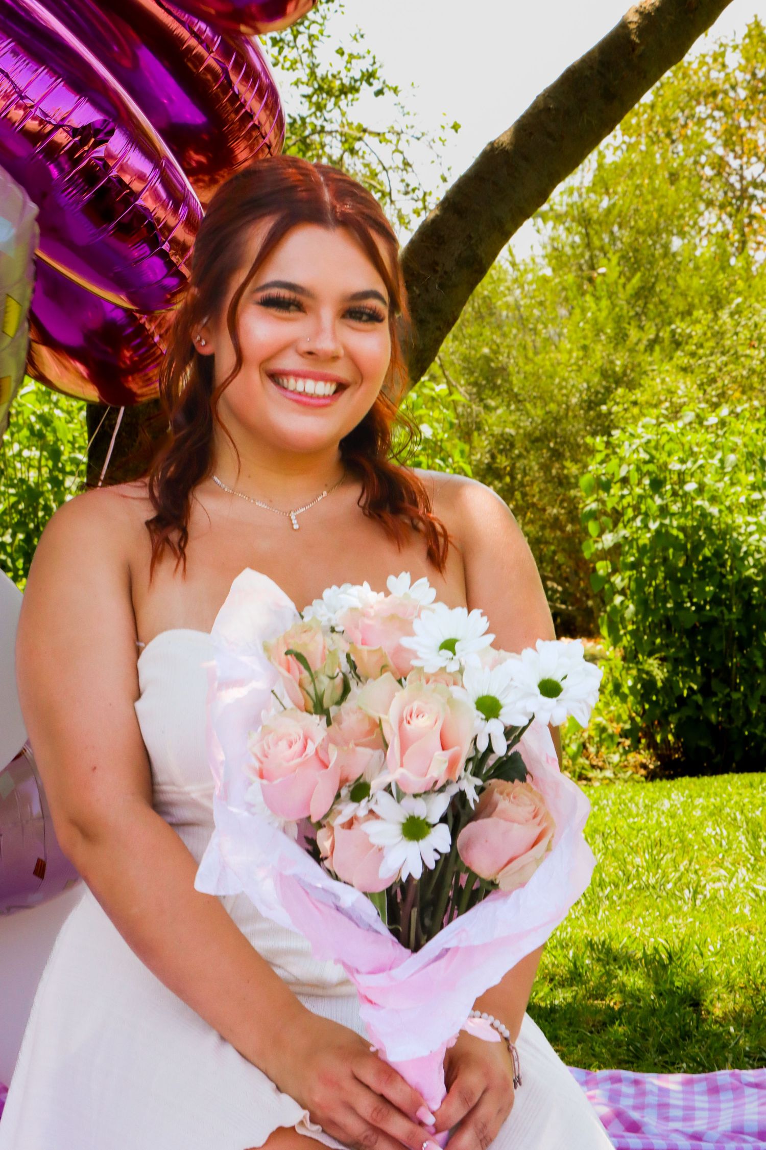 Outdoor celebration with pink roses, white dress, and colorful balloons against green foliage.