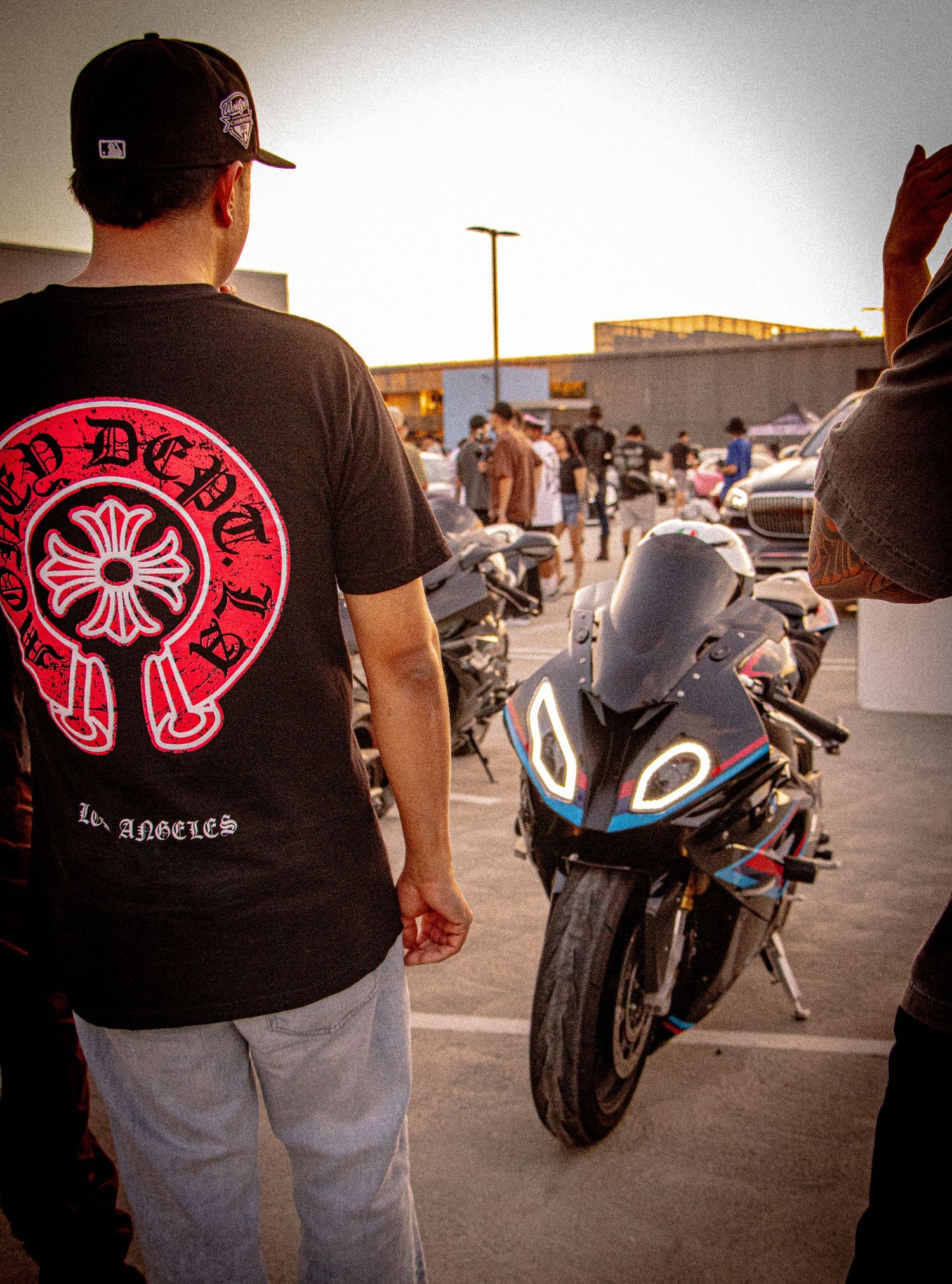 Man in Chrome Hearts LA t-shirt stands beside a sport motorcycle at an outdoor car meet in Los Angeles.