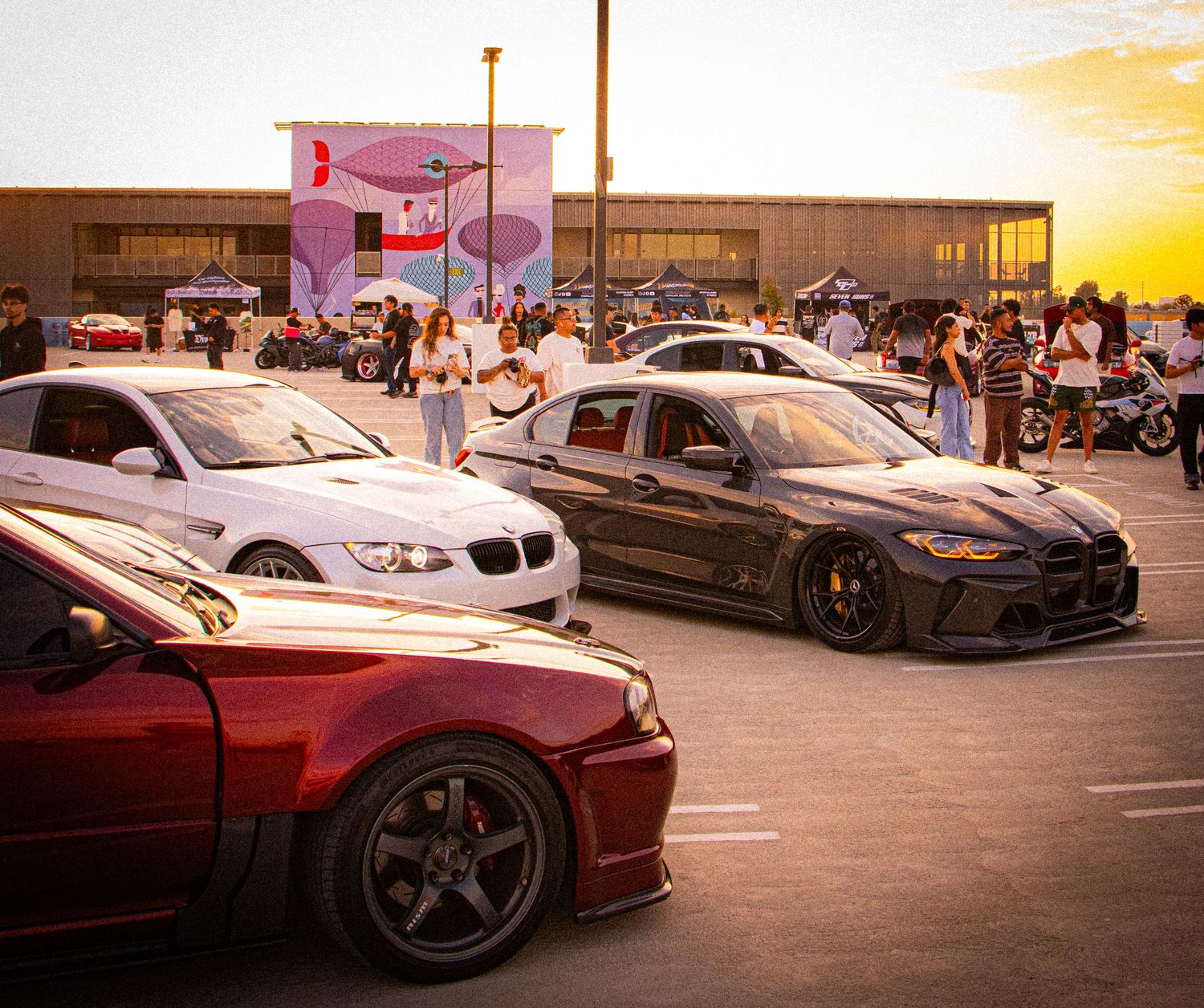 Modified BMW cars displayed at a sunset car show in a parking lot with spectators in the background.