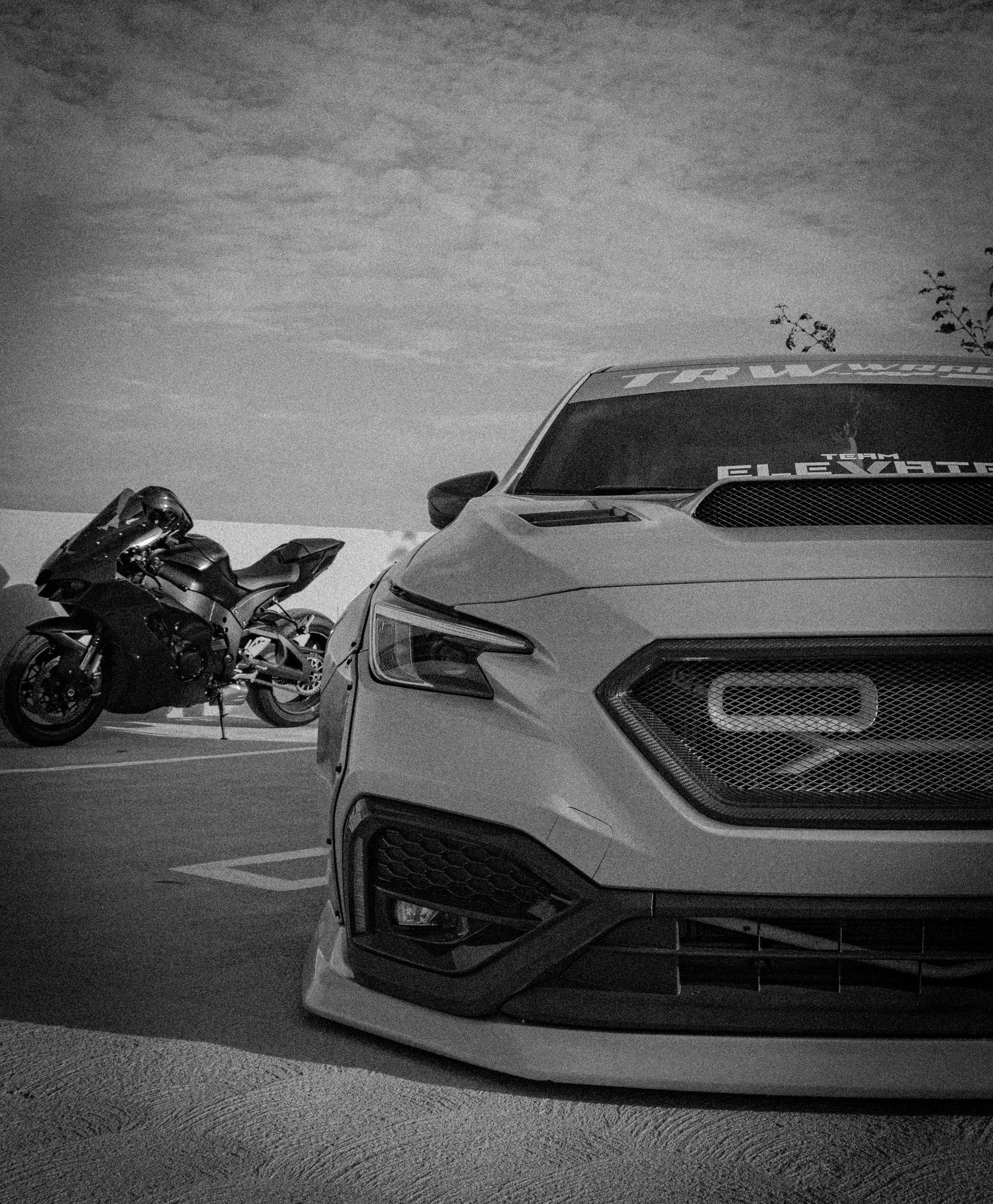 Black and white photo of a modified Subaru WRX with aggressive body kit parked near a sport motorcycle in a parking lot.