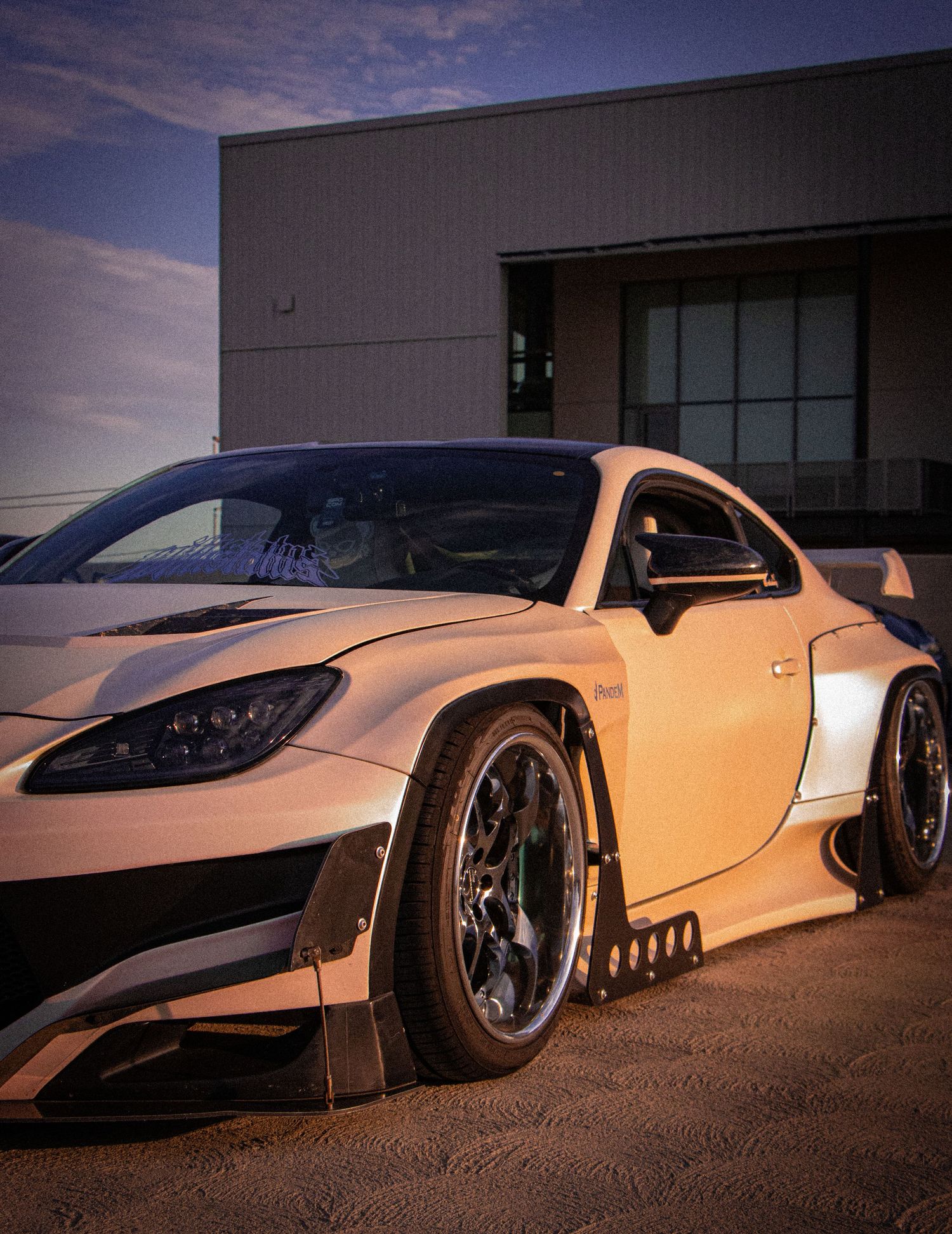 White widebody sports car with aggressive aero kit and dark wheels parked outside a modern building at dusk.