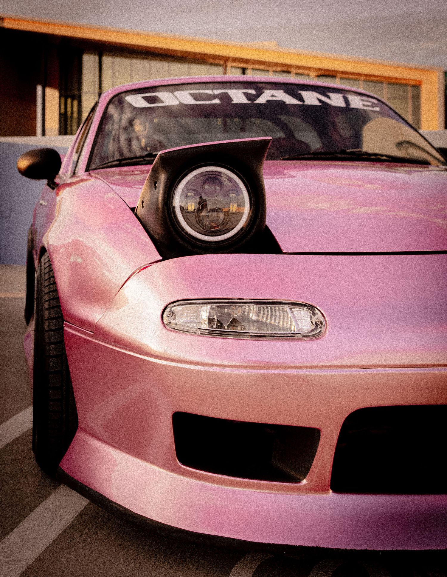 Pink Mazda Miata with pop-up headlights and Octane windshield banner parked outside a building.