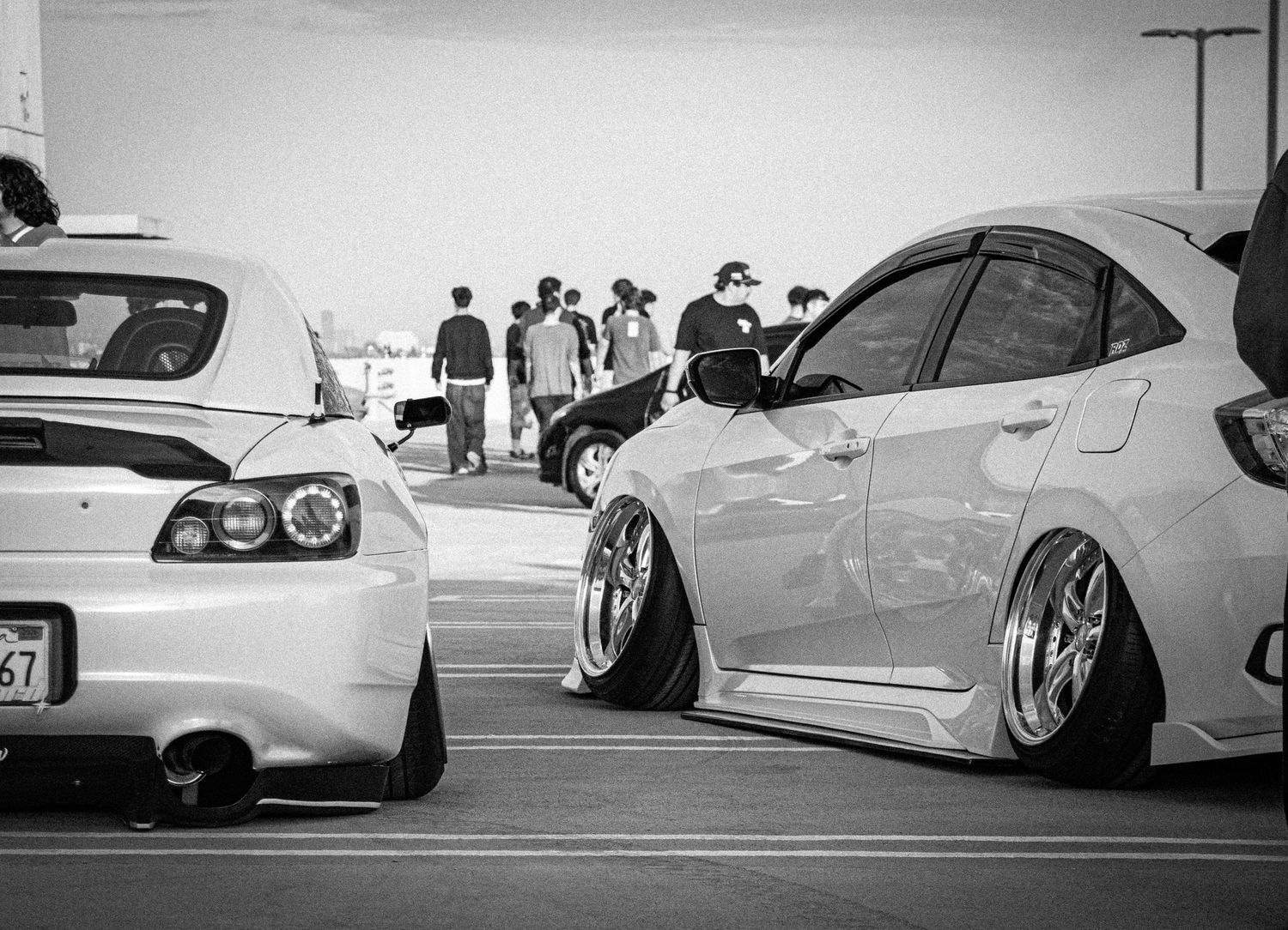 Black and white photo of lowered custom cars at a car meet with spectators gathered in the background.