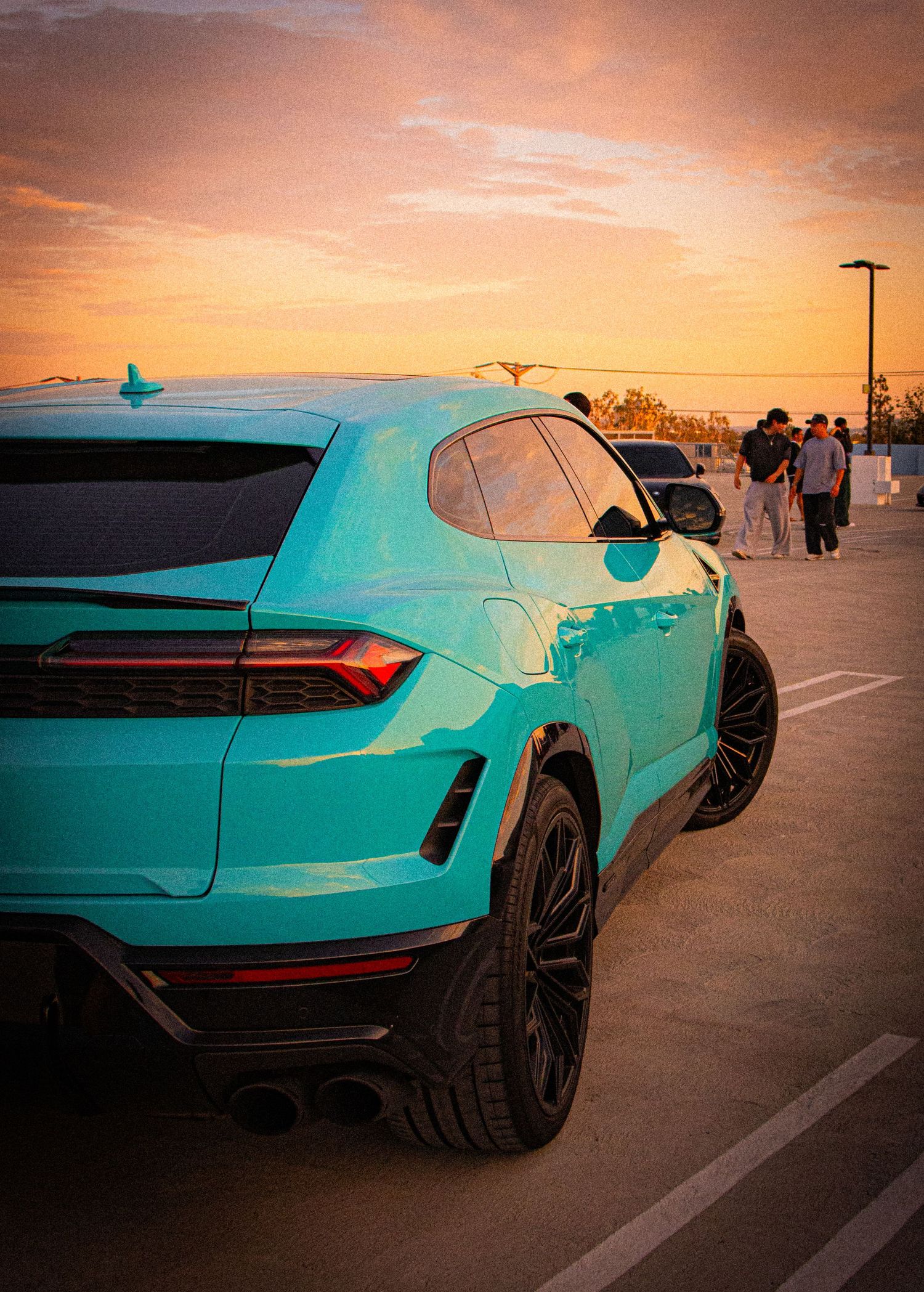 A teal Lamborghini Urus SUV parked at sunset with a vibrant orange sky backdrop in a parking lot.