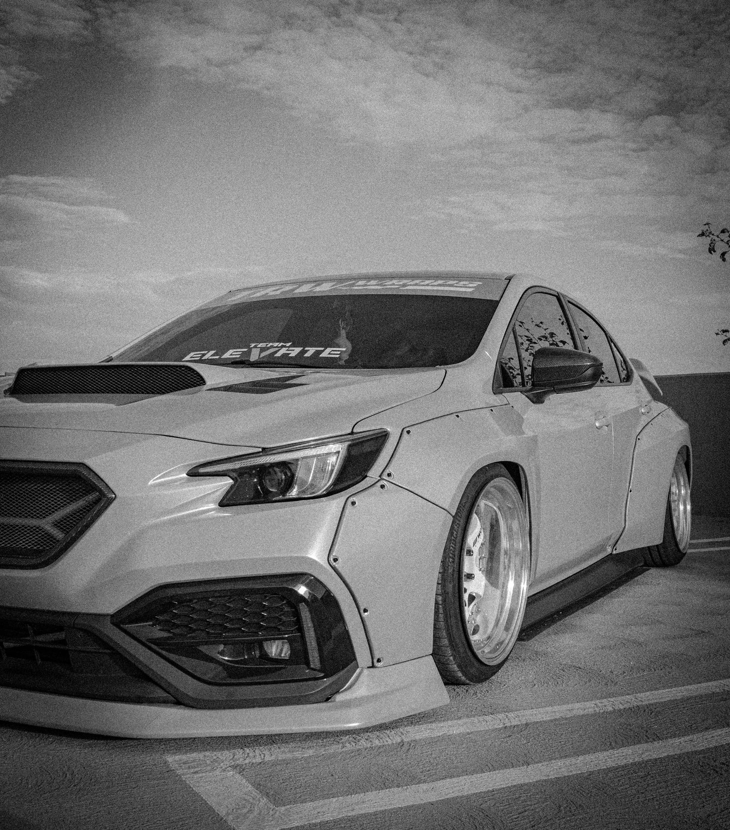 Black and white photo of a modified Subaru WRX with aggressive body kit, lowered stance, and custom wheels in a parking lot.