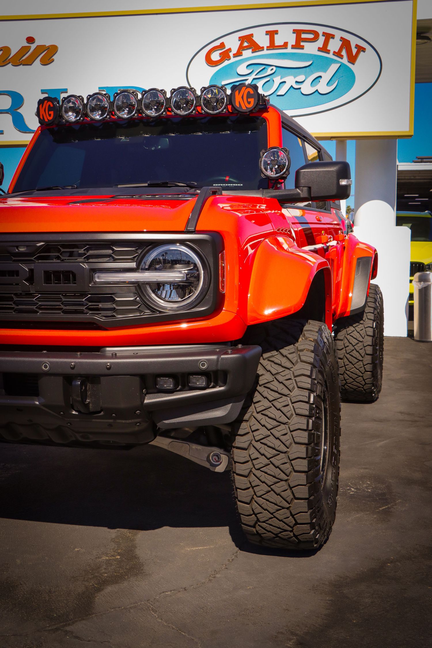 Red Ford Bronco with off-road modifications and roof-mounted light bar at a dealership.