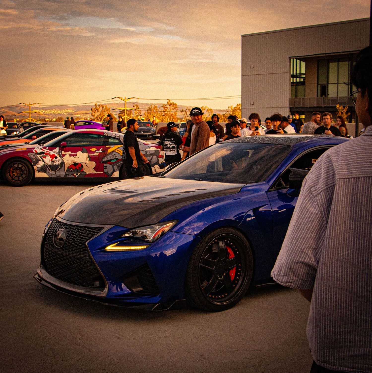 A blue Lexus RC F with carbon fiber accents and red brake calipers at a car meet during golden hour sunset.