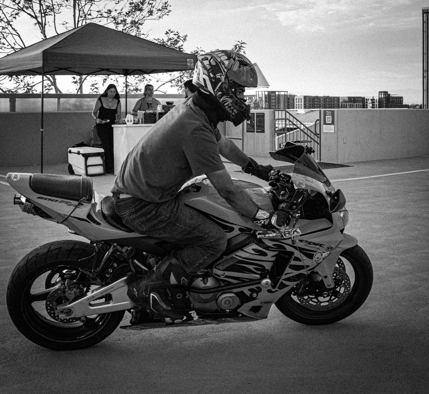 Black and white photo of a motorcyclist in helmet riding a sport bike at an outdoor event with a canopy tent in background.