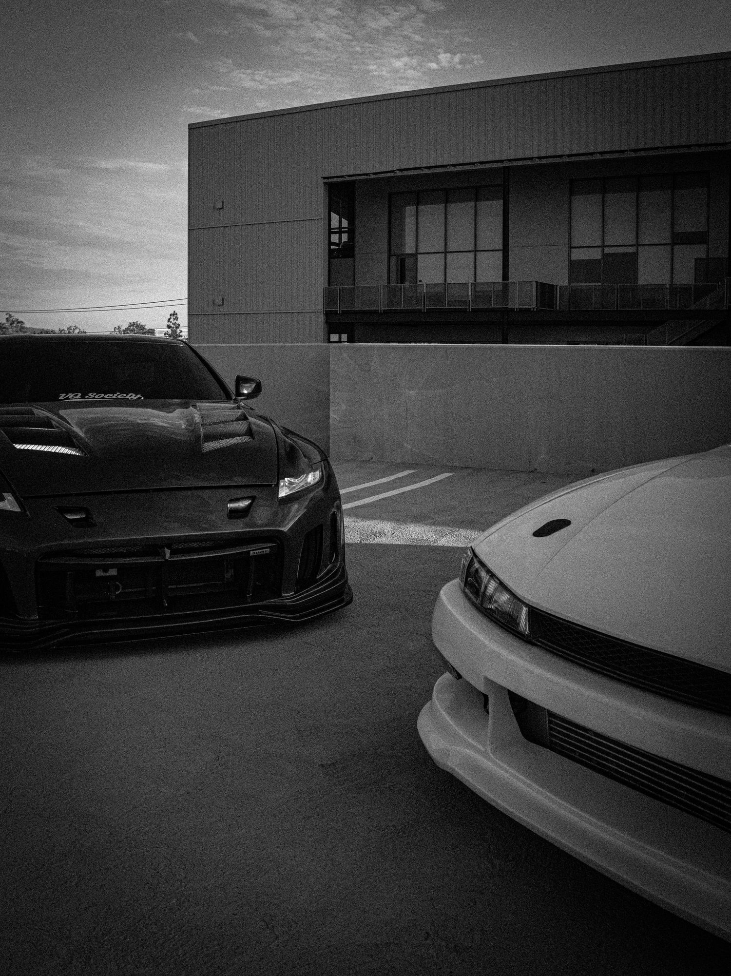 Two modified JDM sports cars, one black and one white, parked outside a building at dusk in a dramatic black and white photo.