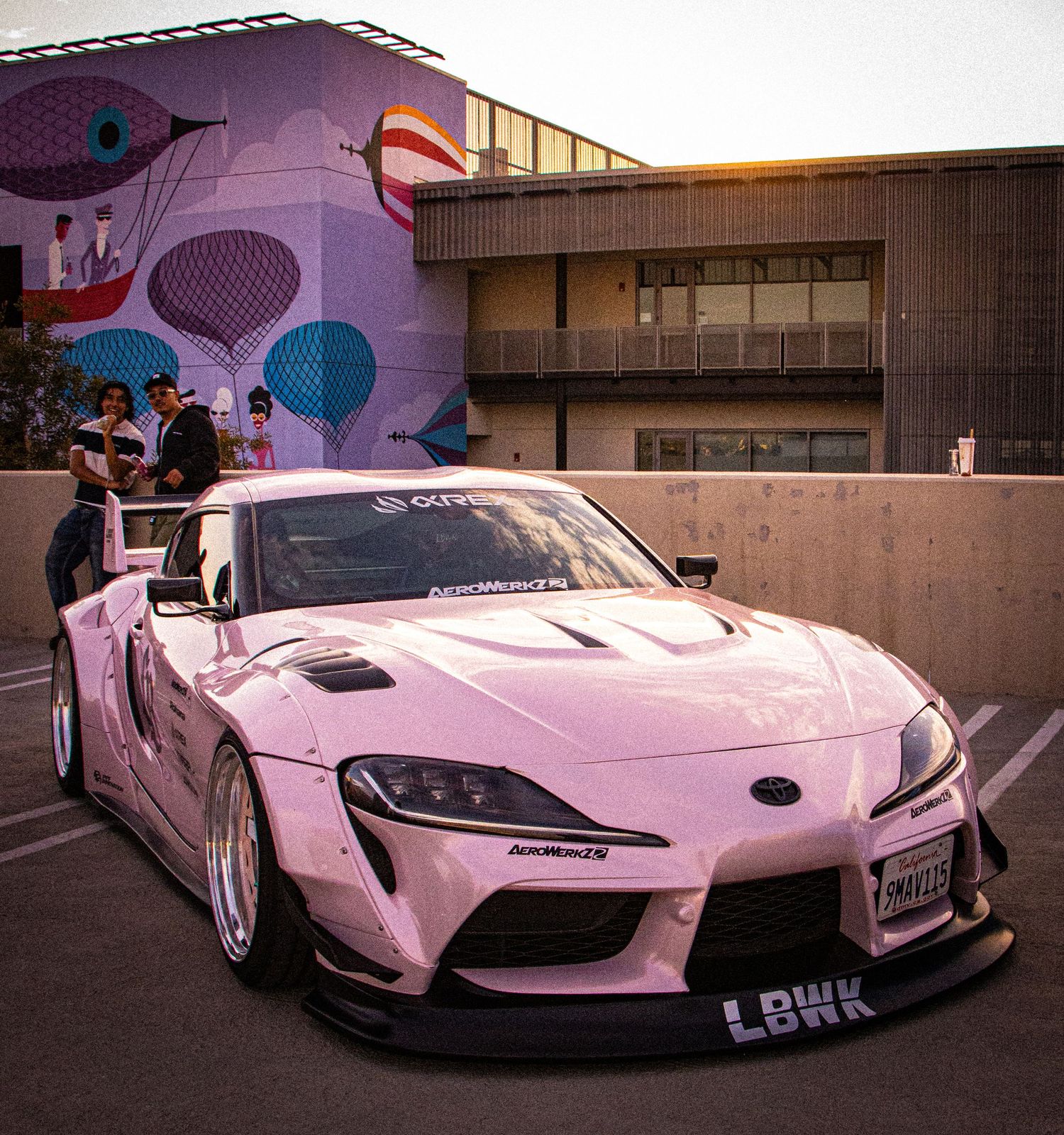 Pink widebody Toyota Supra GR with custom bodykit parked at a car show with colorful mural backdrop.