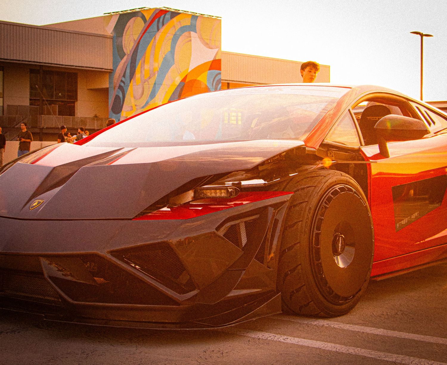 A sleek black Lamborghini Gallardo supercar parked outdoors at golden hour with a colorful mural in the background.