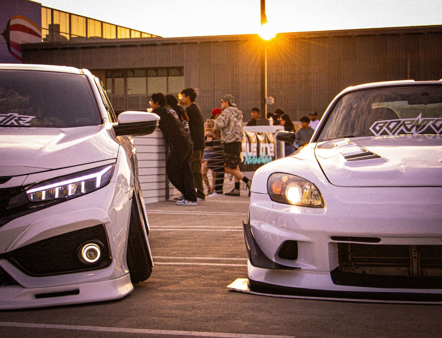 Two white modified Honda cars parked at a sunset car meet with spectators gathered in the background.