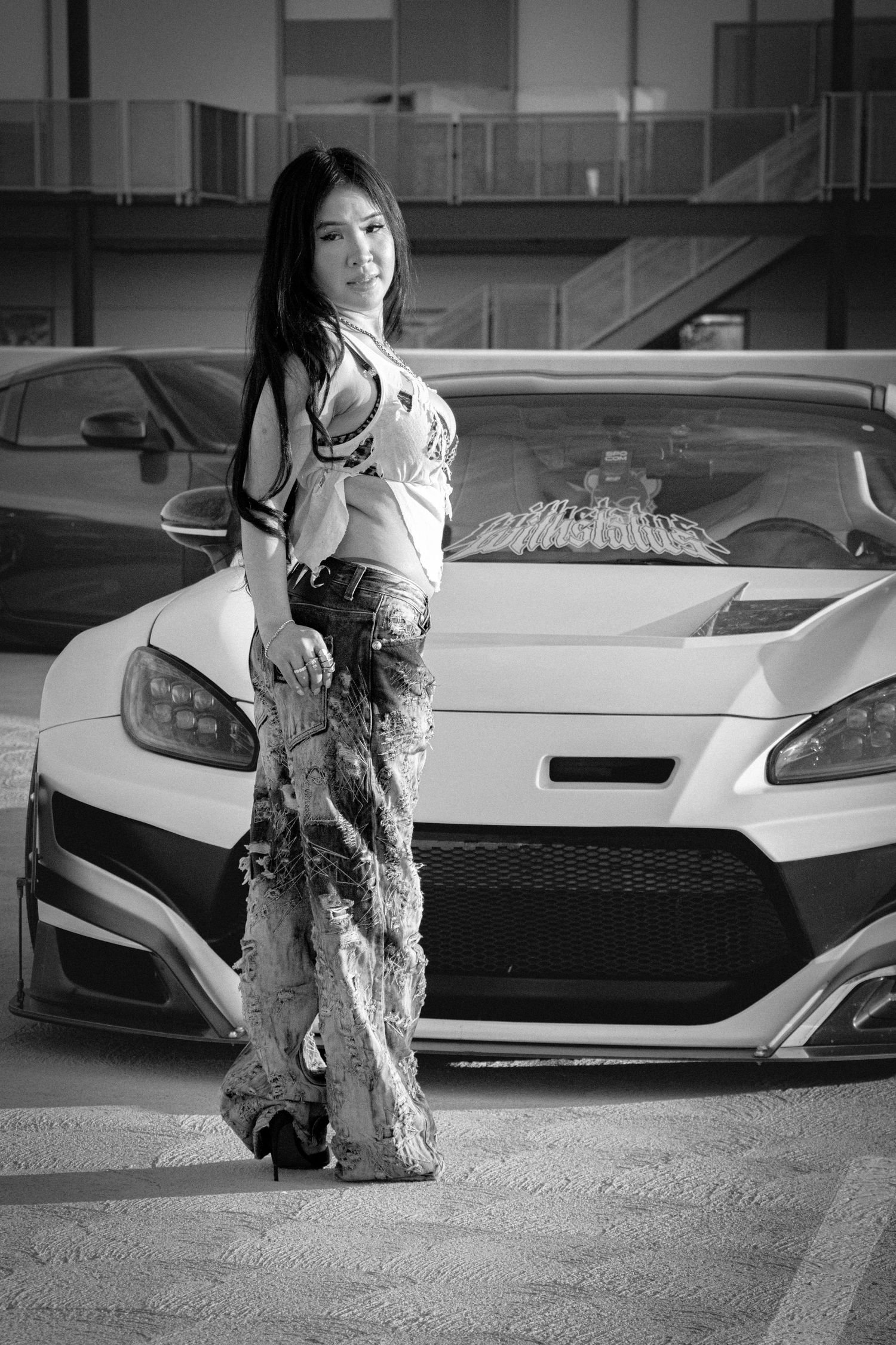 Black and white photo of a woman in stylish outfit posing in front of a sports car in a parking garage.