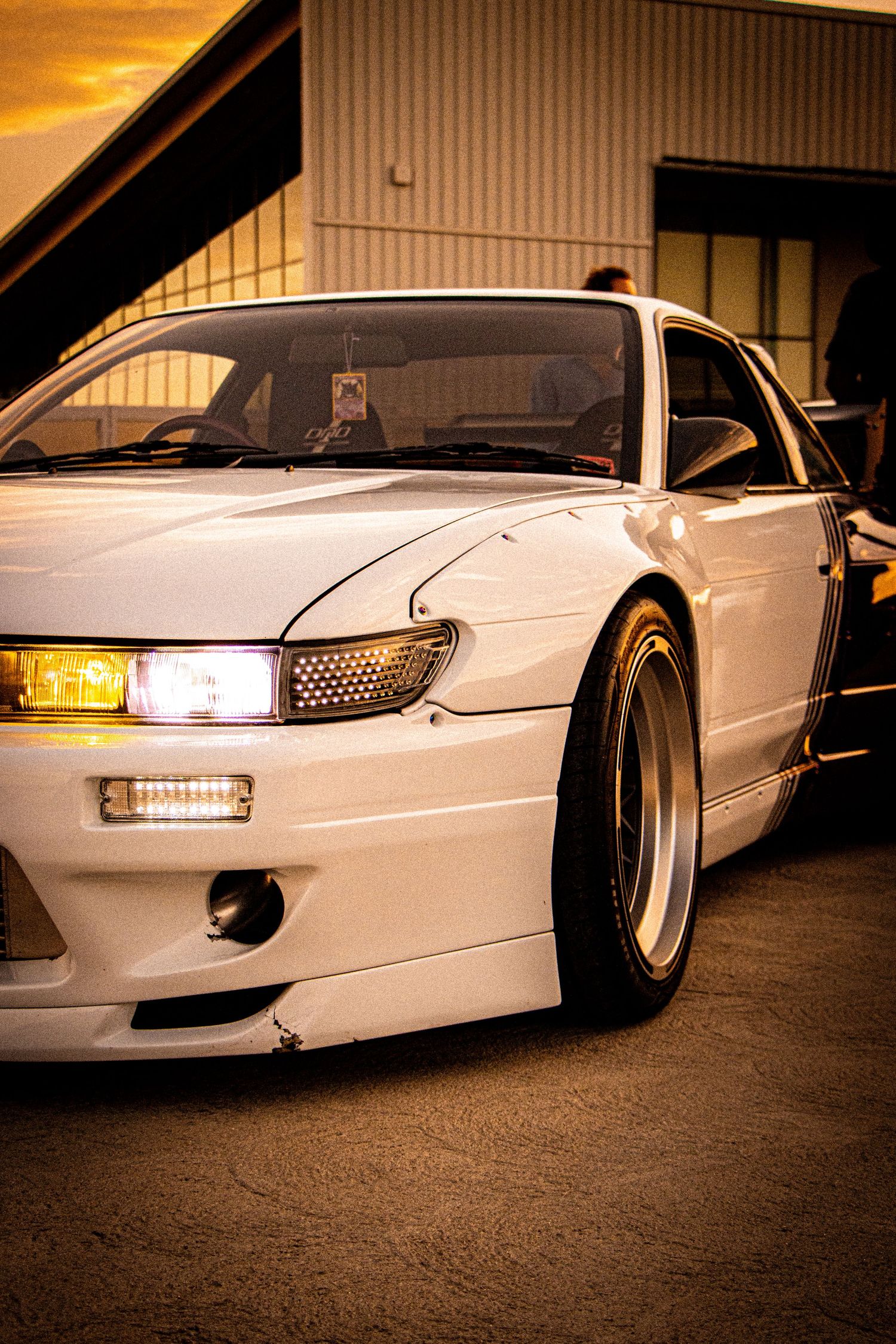 White JDM Nissan Silvia S13 with yellow headlights and wide body kit parked at sunset golden hour.