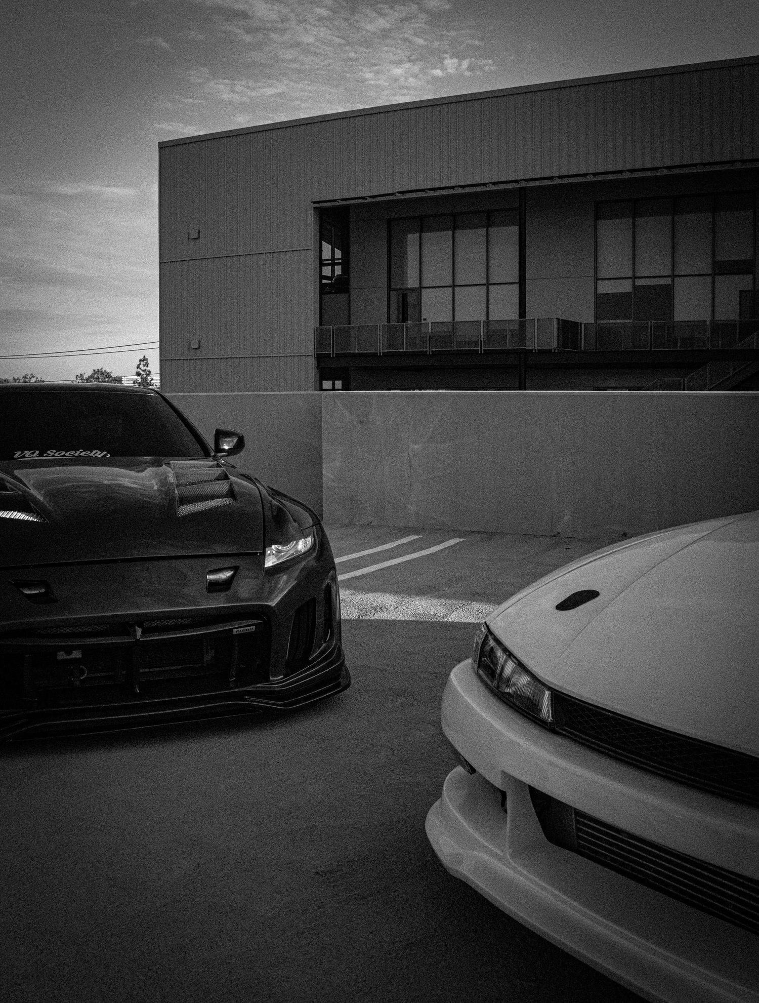 Two modified JDM sports cars, a black and white, parked in a lot beside a modern building at dusk.
