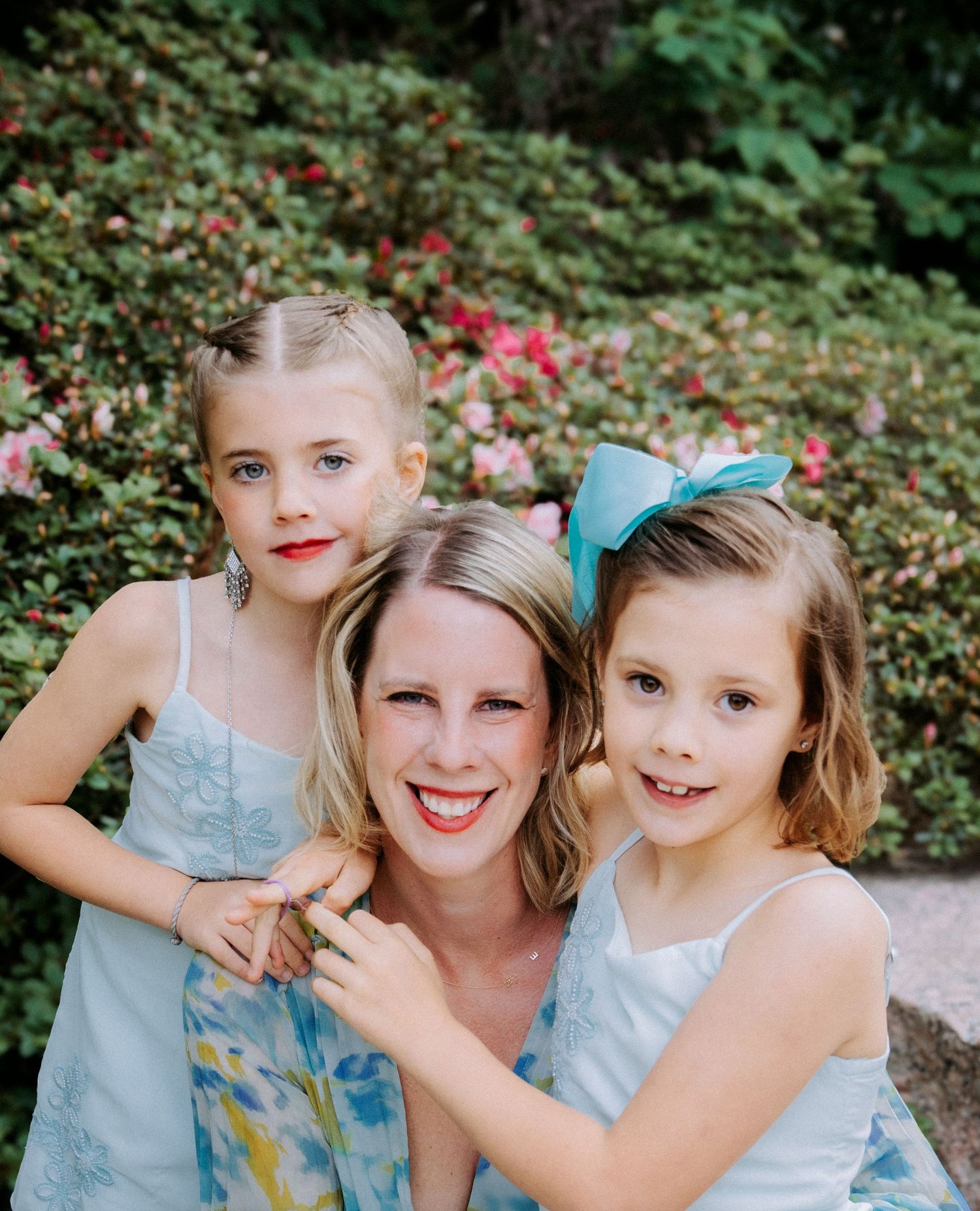 A smiling woman poses outdoors with two young girls in matching light blue dresses, surrounded by lush green shrubs and pink flowers.