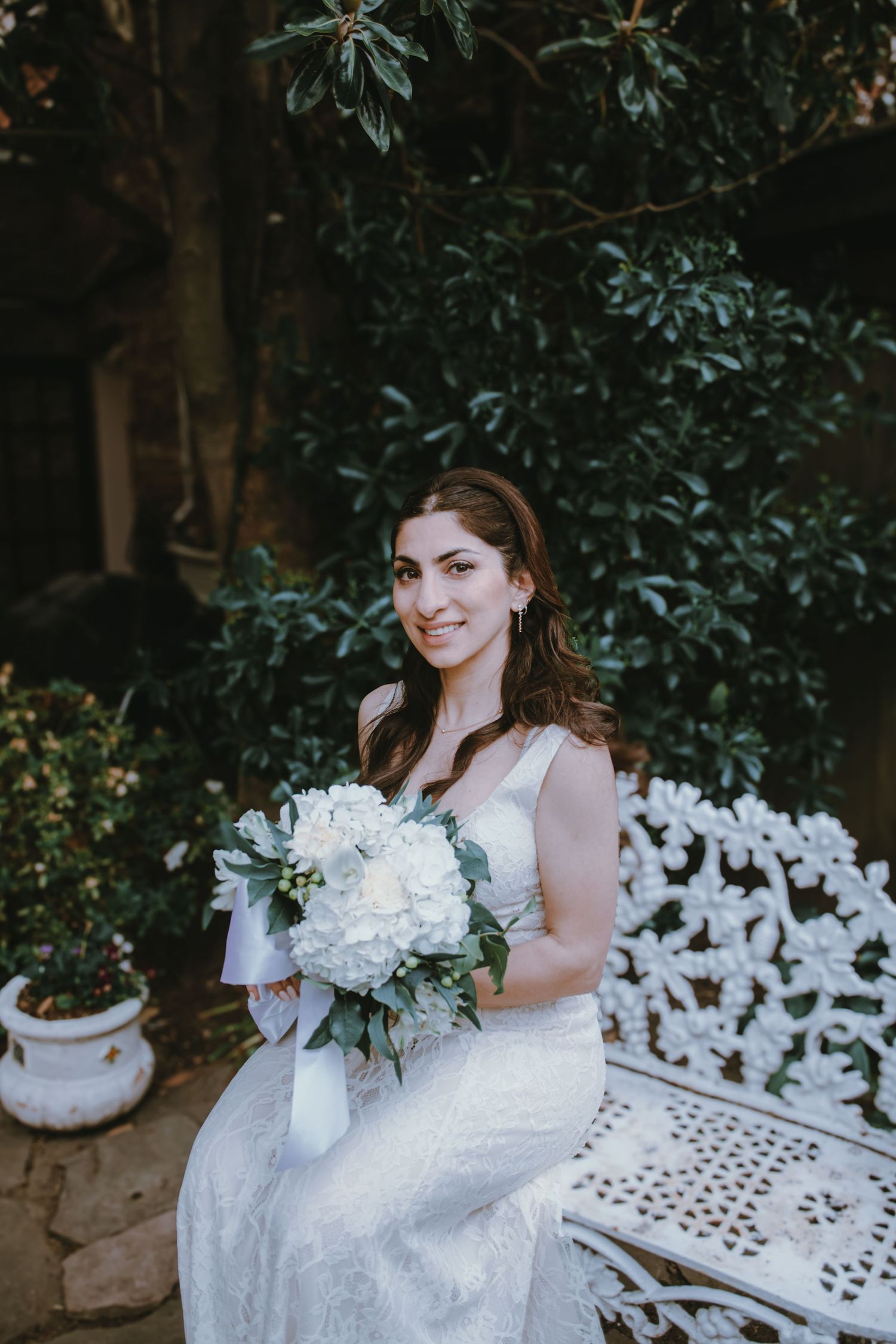 Bride in white lace gown sits on ornate bench holding white hydrangea bouquet, smiling in lush garden setting.