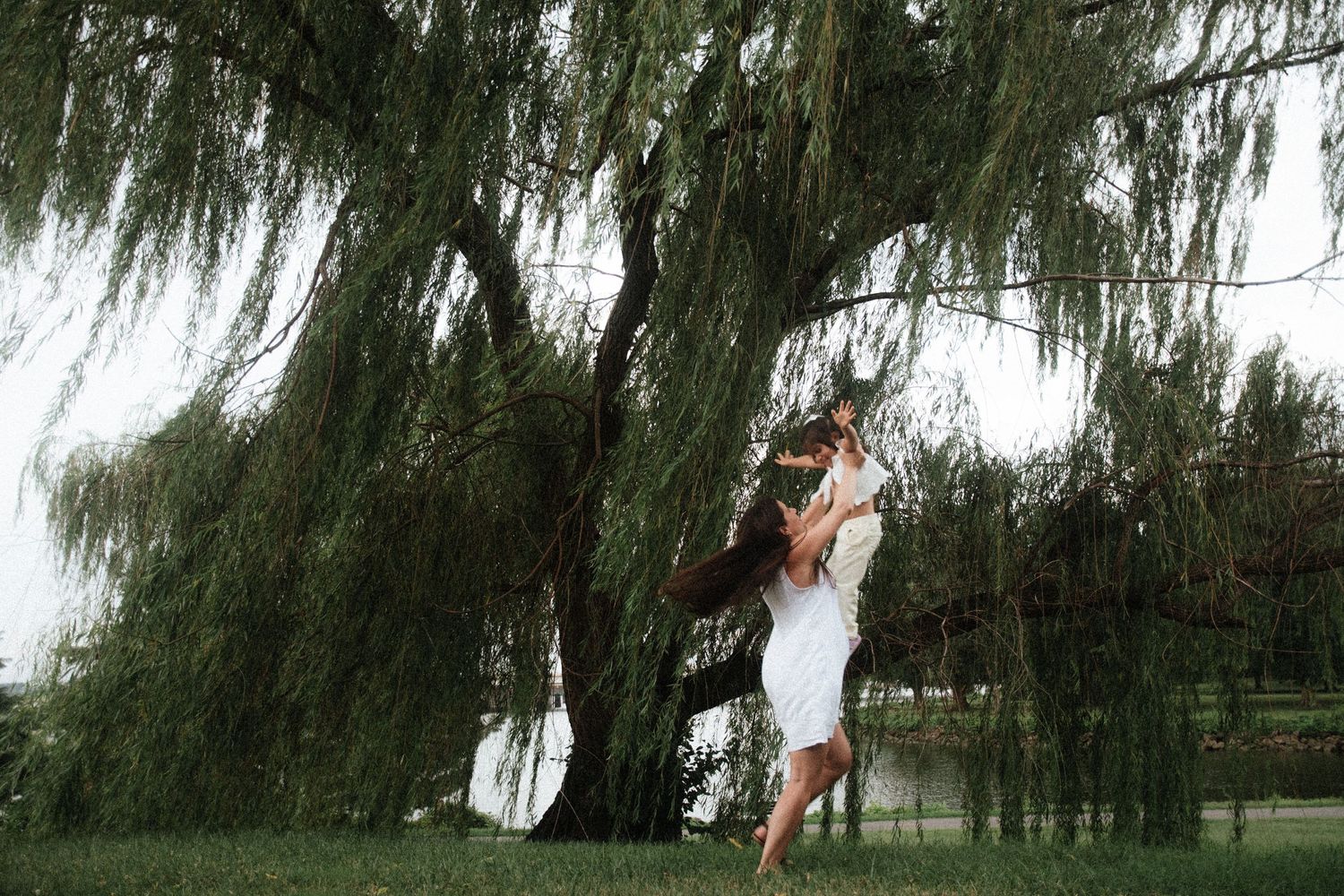 Couple embracing romantically under a weeping willow tree by a lake, woman in white dress lifted by partner.