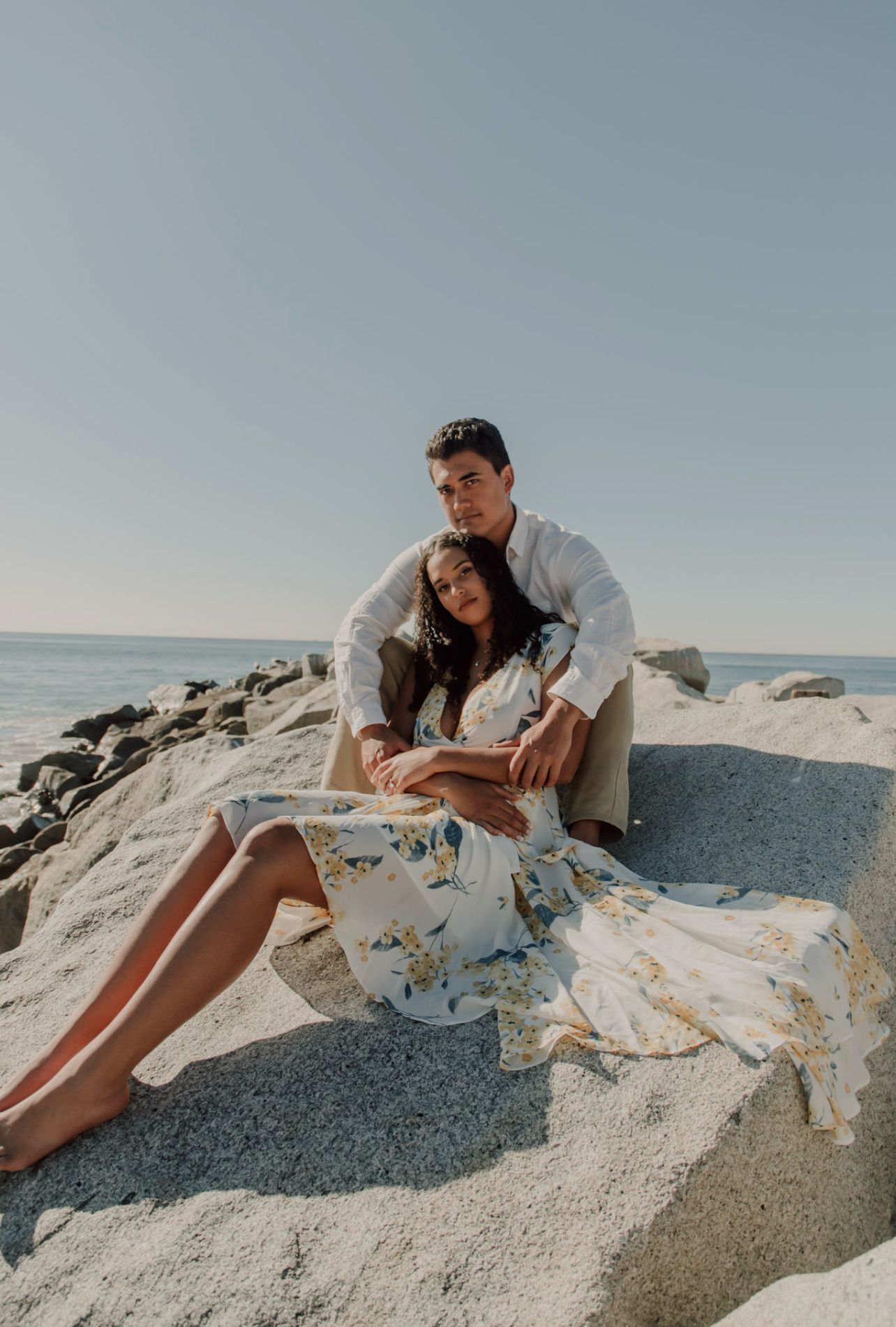 Couple relaxes on rocky beach during sunny outdoor photoshoot.
