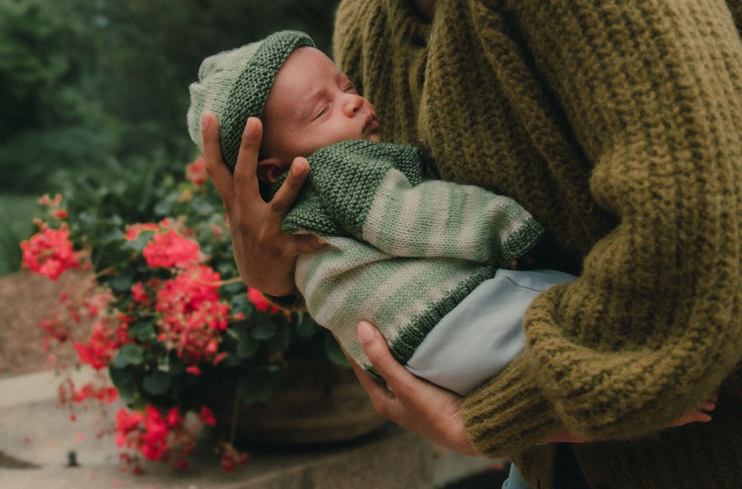 Adult cradling a sleeping newborn wrapped in green knit blanket and hat near red flowers outdoors.