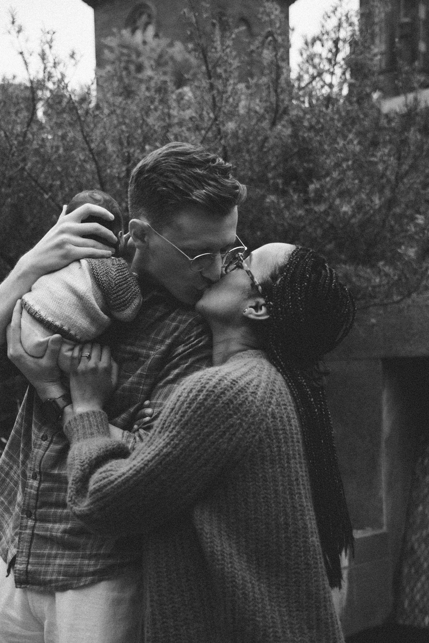Black and white photo of a couple kissing while holding their baby outdoors near lush greenery.