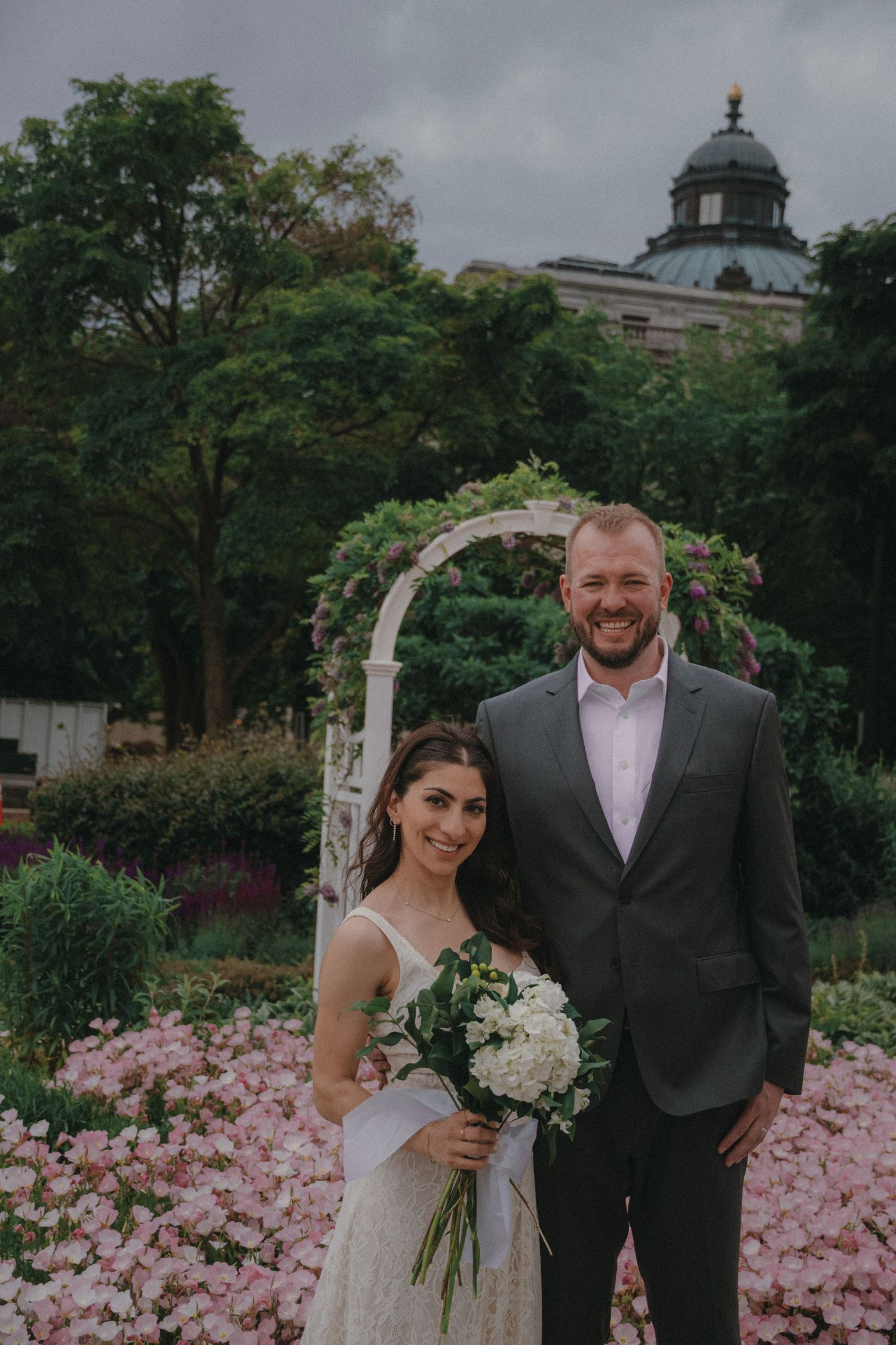 Couple poses in garden with white floral arch, pink flowers, and historic building backdrop on wedding day.