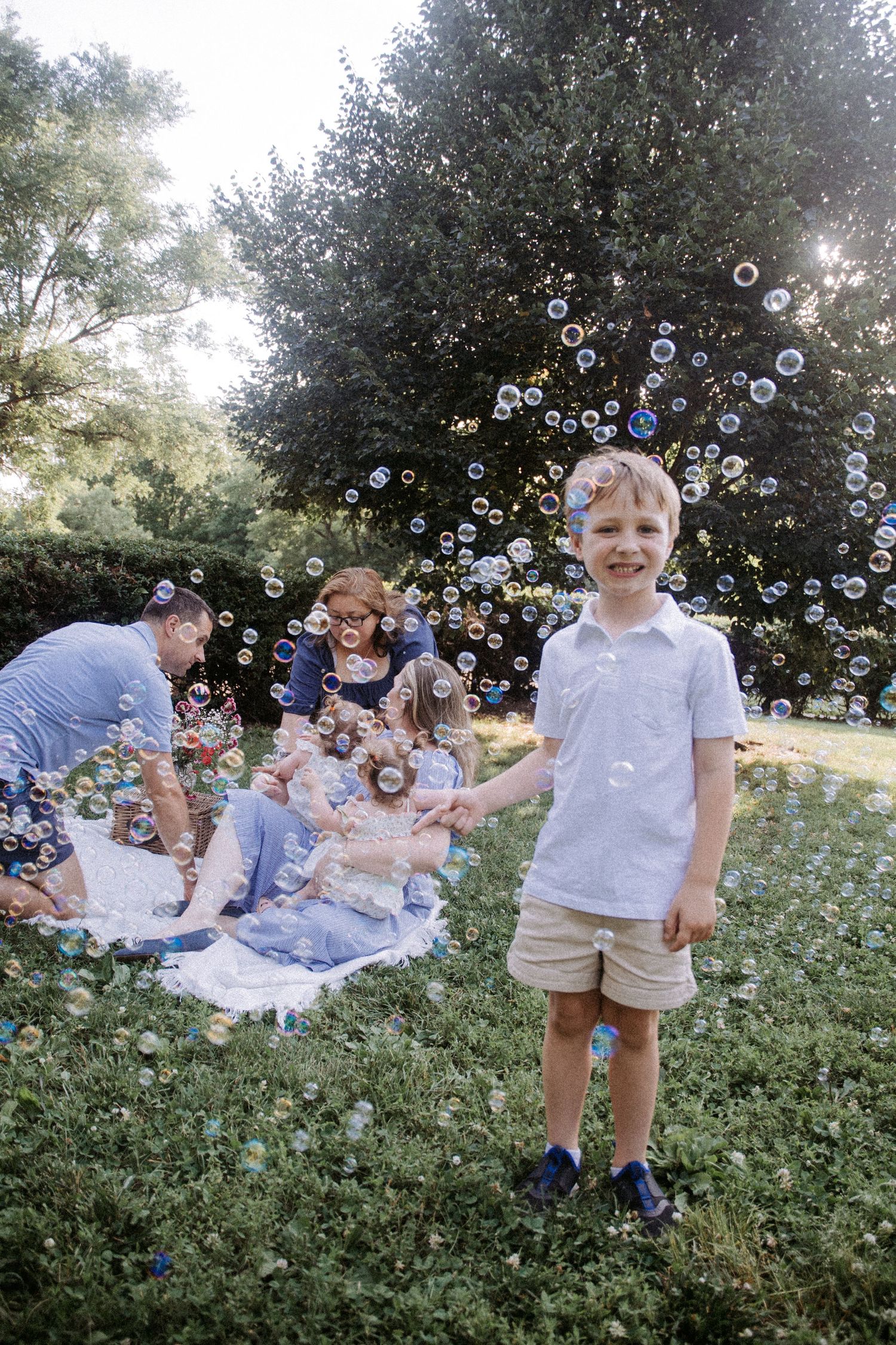 Young boy smiling outdoors surrounded by soap bubbles, family enjoying a picnic on grass in a lush green park.