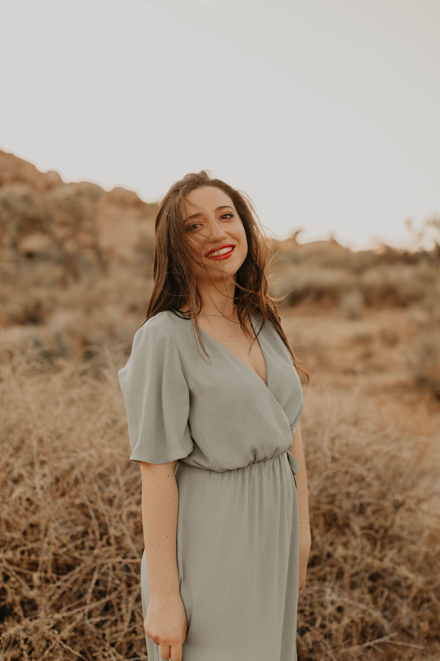 A woman in a sage green dress poses in a desert landscape during golden hour.